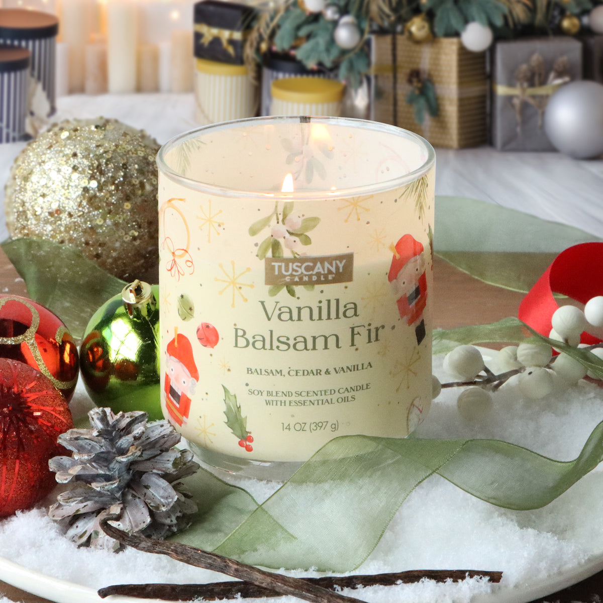 A lit Tuscany Candle® SEASONAL Vanilla Balsam Fir: Cedar & Vanilla Scented Holiday Candle (14 oz) in a glass jar, surrounded by snow, pinecones, ornaments, and ribbons.