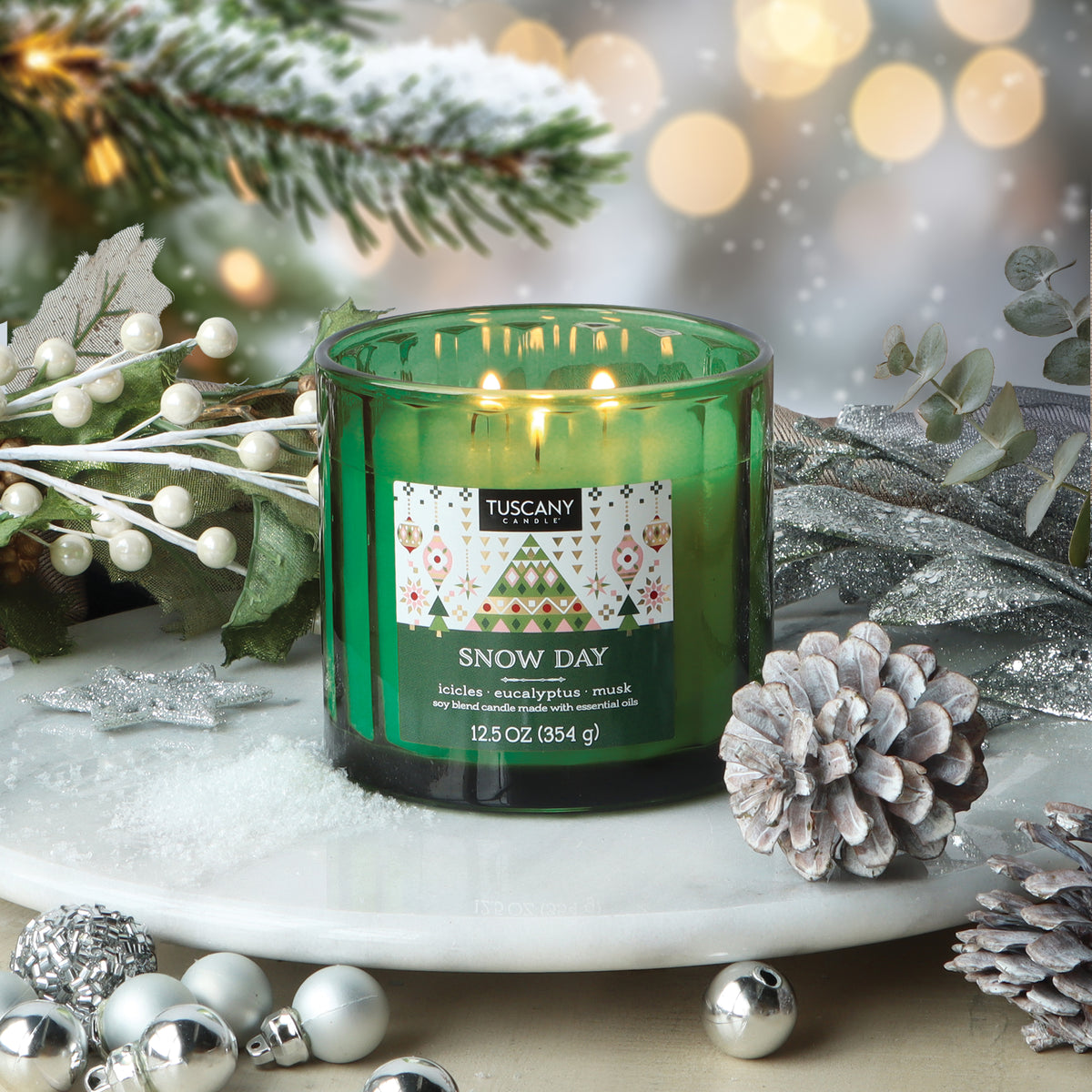 A Tuscany Candle® SEASONAL Snow Day: Eucalyptus & Musk Scented Holiday Candle (12.5 oz) in green glass sits on a marble tray surrounded by pinecones, silver ornaments, and snowy greenery.