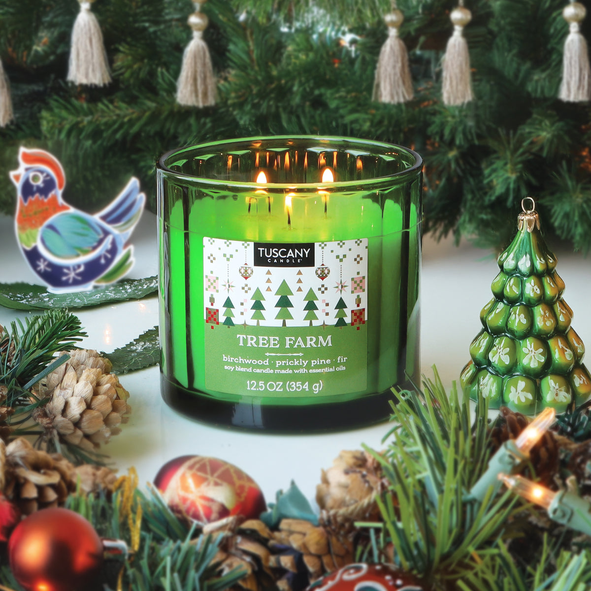 A green Tuscany Candle® SEASONAL Tree Farm: Warm Birchwood & Pine Scented Holiday Candle (12.5 oz) in a glass jar, surrounded by Christmas ornaments, pinecones, and evergreen branches.