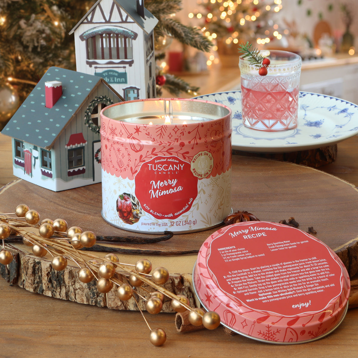 Merry Mimosa: Pomegranate & Rhubarb Scented Holiday Candle (12 oz) in a pink tin with a cocktail recipe lid, set on a wooden surface next to a glass of pink drink.