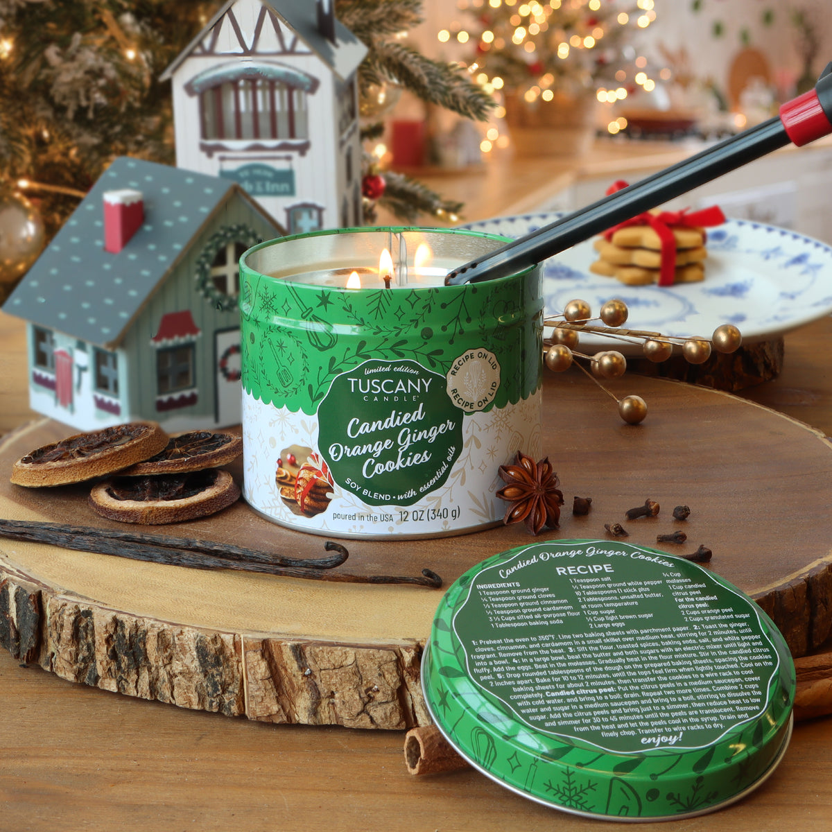 A Tuscany Candle® SEASONAL Candied Orange Ginger Cookies Scented Holiday Candle (12 oz) in a green tin with its recipe lid, set on wood with dried orange slices, star anise, and a vanilla bean.