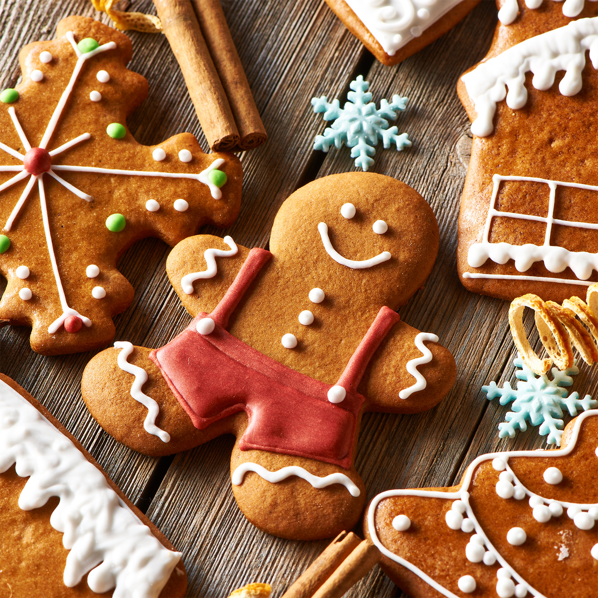 A Candied Orange Ginger Cookies Scented Holiday Candle (12 oz) by Tuscany Candle® SEASONAL, surrounded by gingerbread cookies with white icing, cinnamon sticks, and snowflake ornaments on wood.