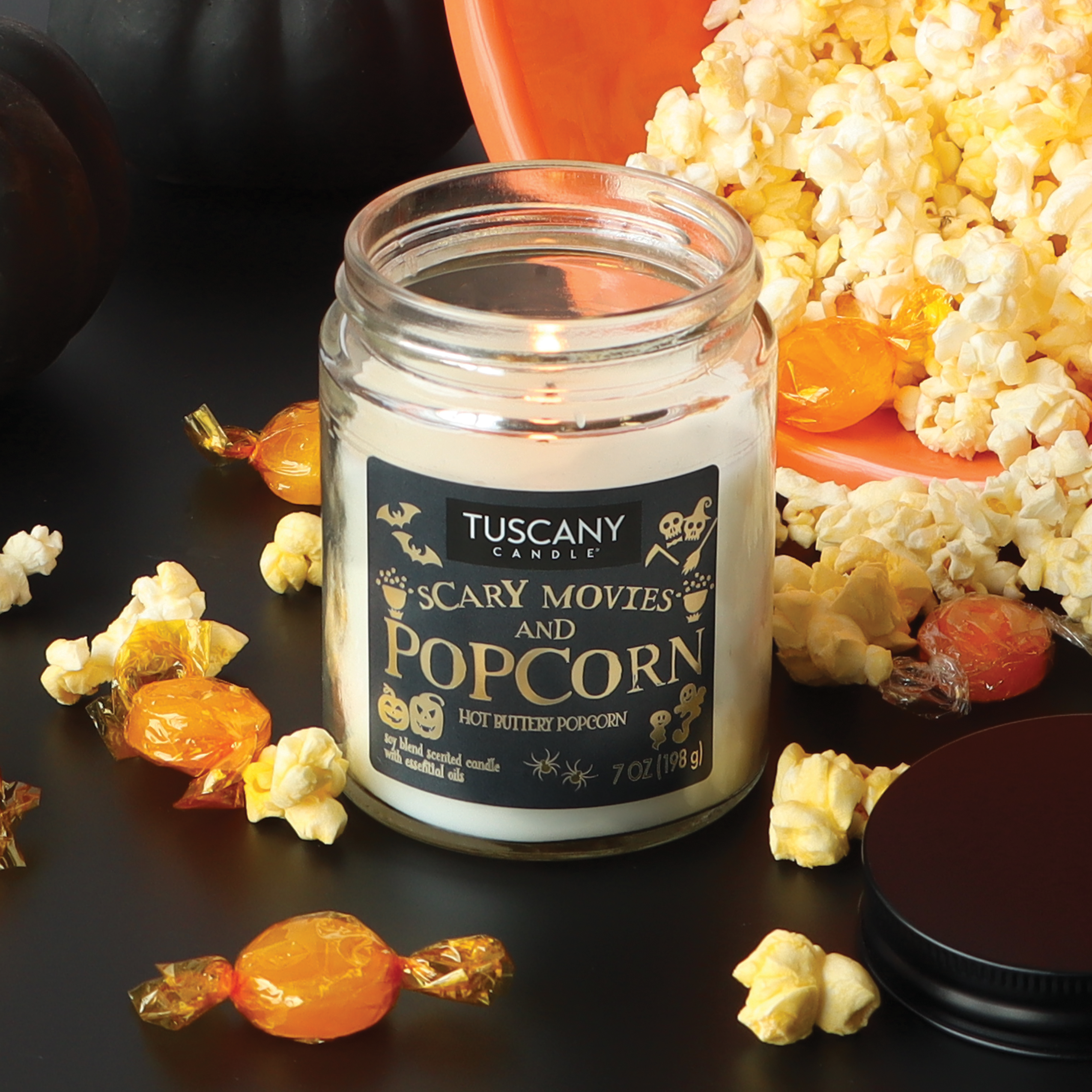 A Tuscany Candle® SEASONAL "Scary Movies & Popcorn" long-lasting scented jar candle (12 oz) casts a warm glow beside a bowl of popcorn and wrapped orange candies on a black surface.