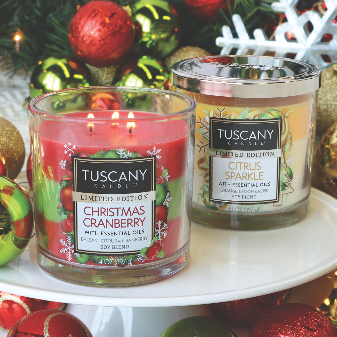 A pair of Tuscany Candle® SEASONAL candles—a "Christmas Cranberry" in red and a yellow "Citrus Sparkle Long-Lasting Scented Jar Candle (14 oz)"—gracefully surrounded by ornaments, adorn the scene before a festively decorated Christmas tree.