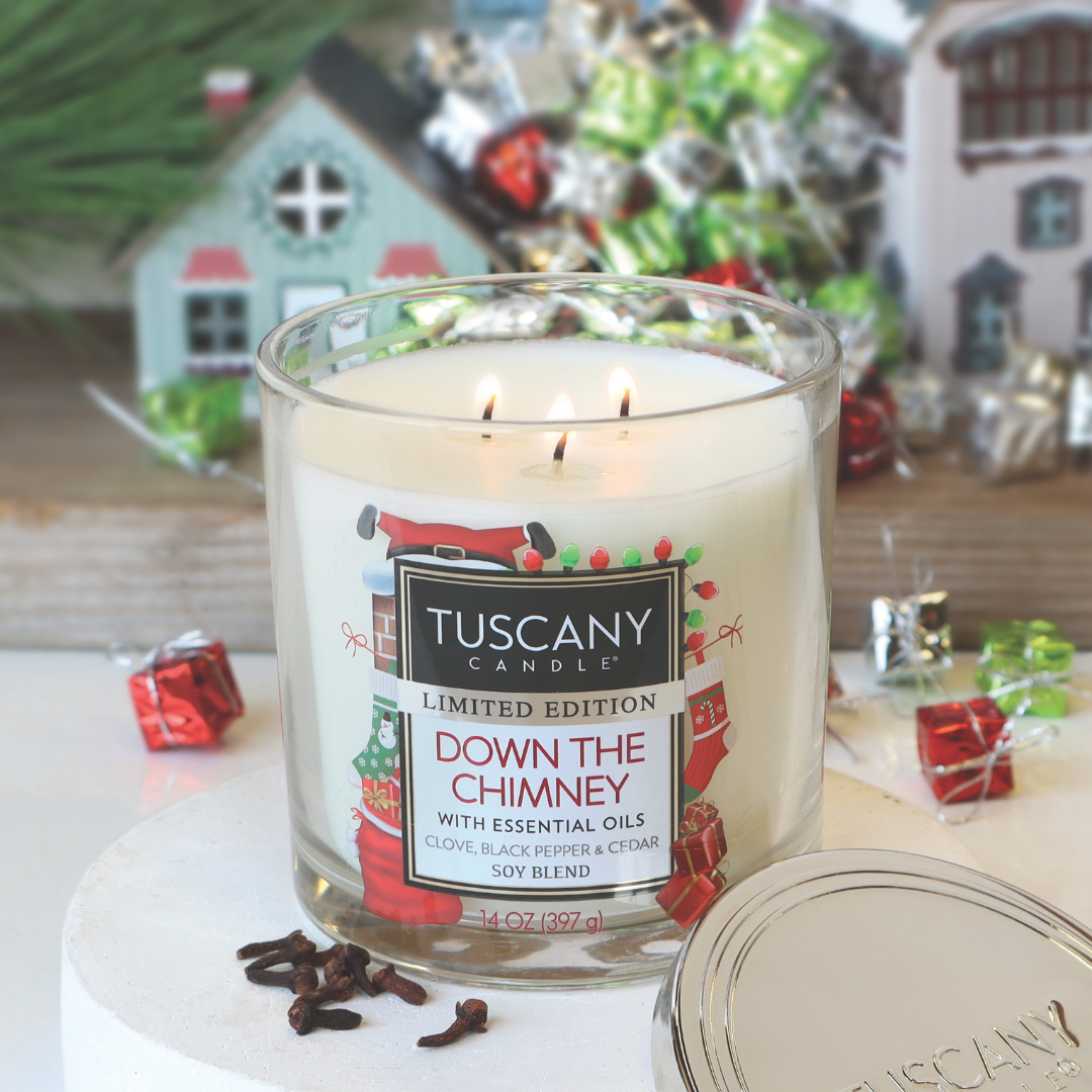 A Tuscany Candle® SEASONAL "Down The Chimney" long-lasting scented jar candle (14 oz), infused with essential oils and featuring the fragrances of clove, black pepper, and cedar, is displayed alongside small festive gift boxes and miniature houses.