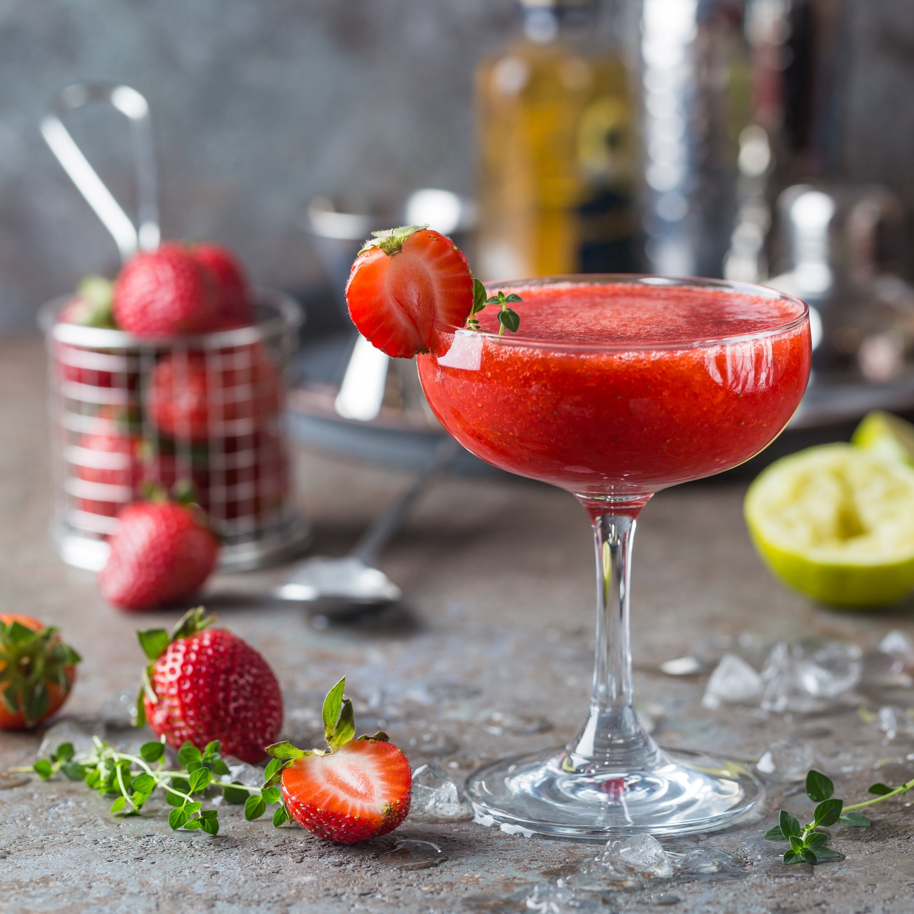 A refreshing *Strawberry Daiquiri (12 oz) – Sunset Beach Bar Collection* garnished with a sliced strawberry and a sprig of thyme is surrounded by fresh strawberries, a halved lime, and some green herbs on a rustic table. *Tuscany Candle® SEASONAL*.
