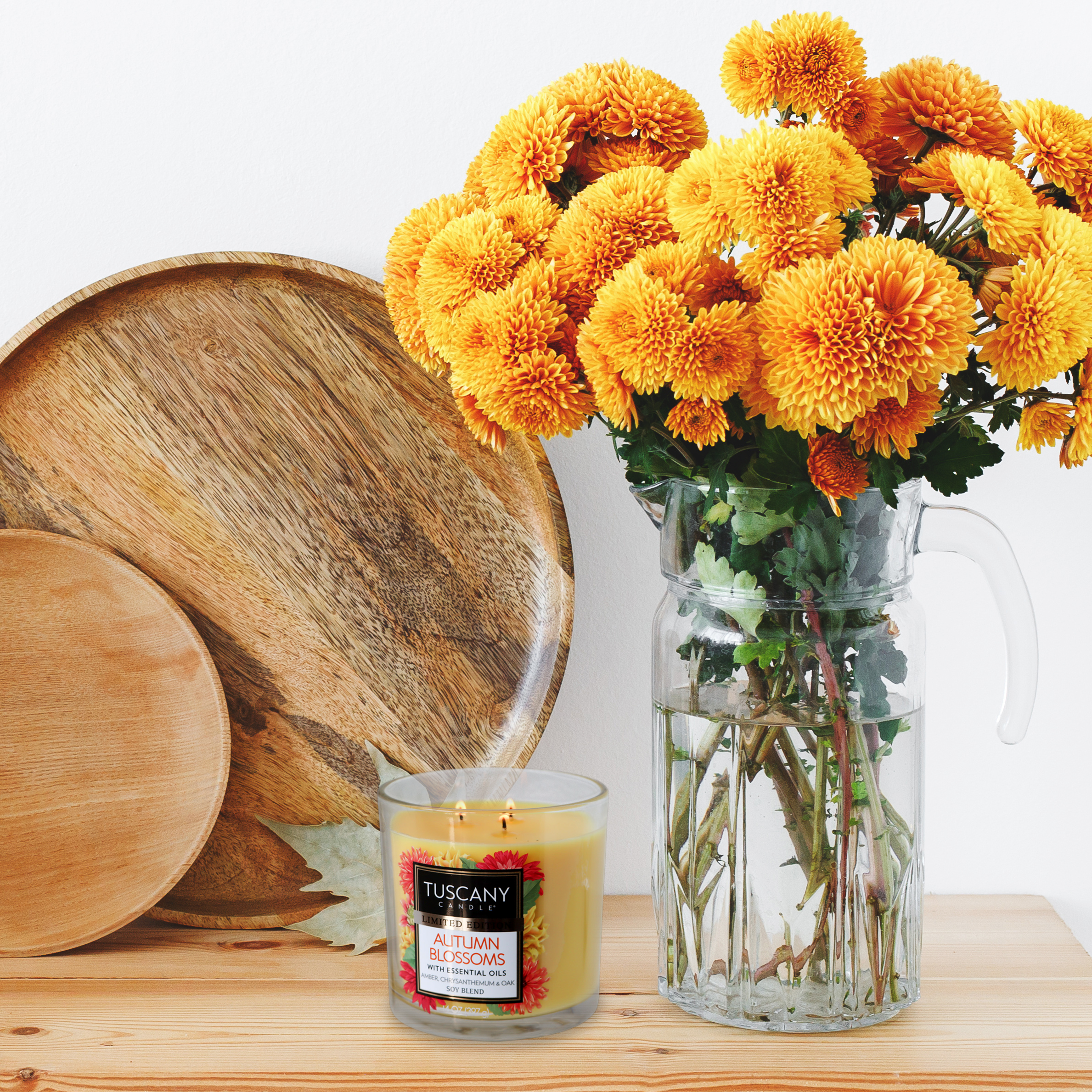 A glass pitcher filled with orange and yellow flowers stands alongside two wooden plates and a lit "Autumn Blossoms Long-Lasting Scented Jar Candle" (14 oz) by Tuscany Candle® SEASONAL on a white surface, exuding an inviting autumn charm.