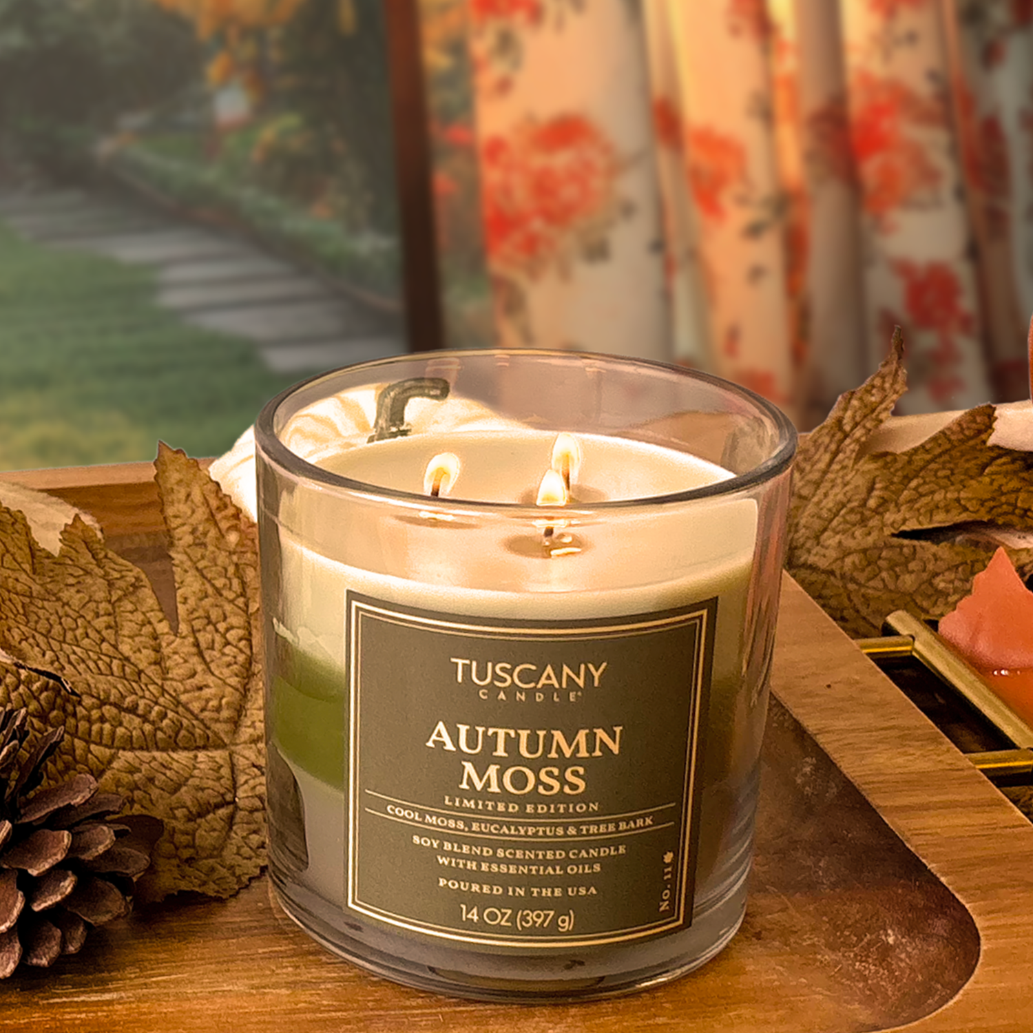 A Tuscany Candle® SEASONAL Autumn Moss: Eucalyptus & Sandalwood Scented Fall Candle (14 oz), made from a soy wax blend with two lit wicks, sits on a tray amid dried leaves and decor, with a blurred curtain and garden in the background.