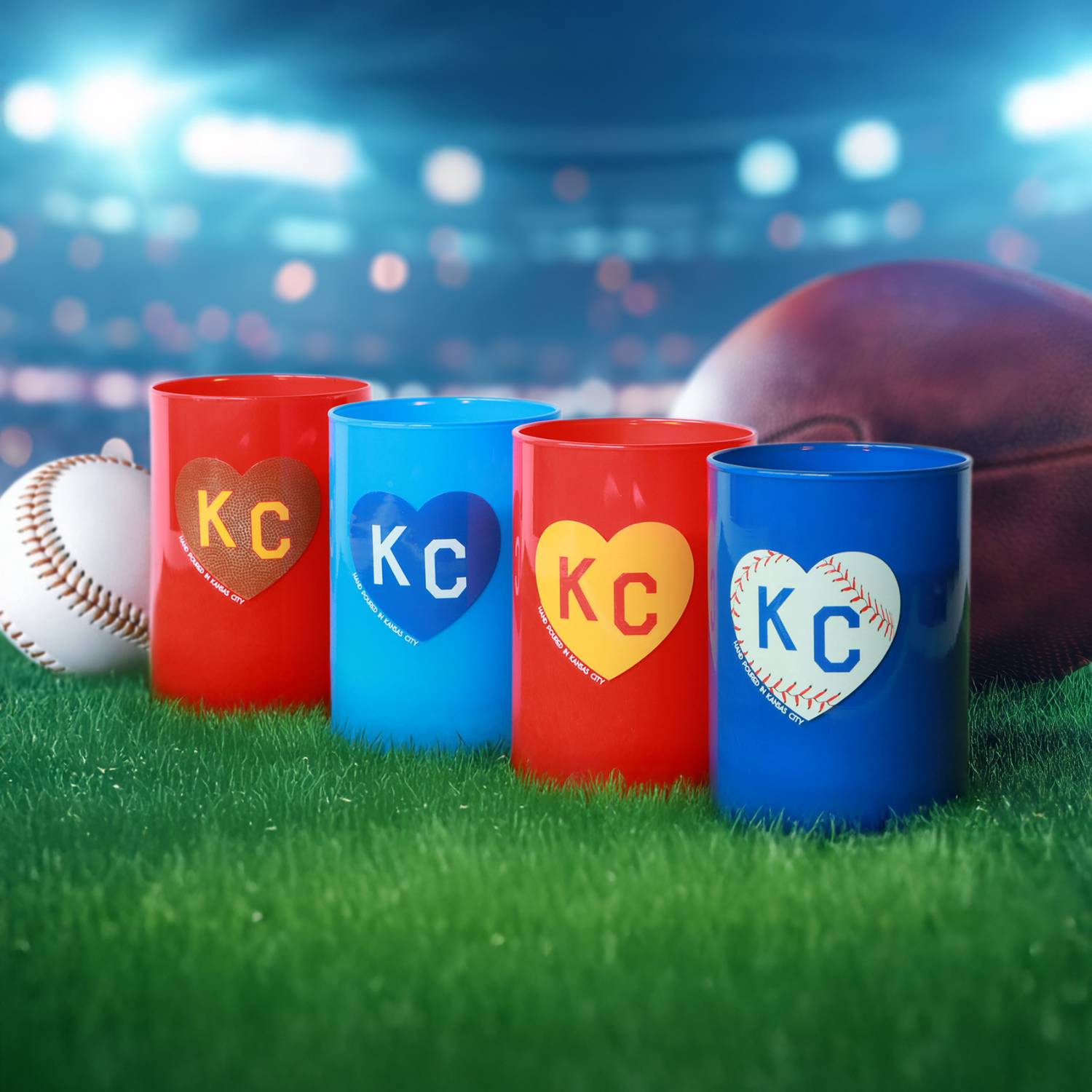 A Fall Festival Scented Red KC Heart Candle (13 oz) by Tuscany Candle x Charlie Hustle sits on grass with a baseball and football, radiating a game day vibe against a blurred stadium backdrop.