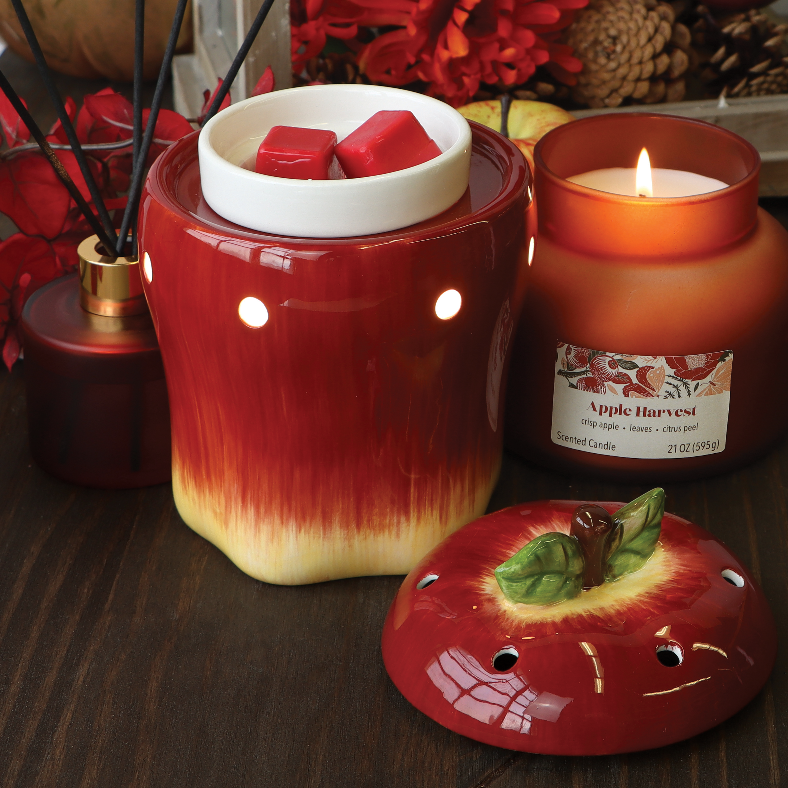 A Tuscany Candle® SEASONAL Apple Wax Melt Warmer, crafted in red ceramic with an apple-shaped lid, sits on a wooden surface next to two wax cubes and a lit "Apple Harvest" scented candle. It's surrounded by reed diffusers and cozy autumn decor.