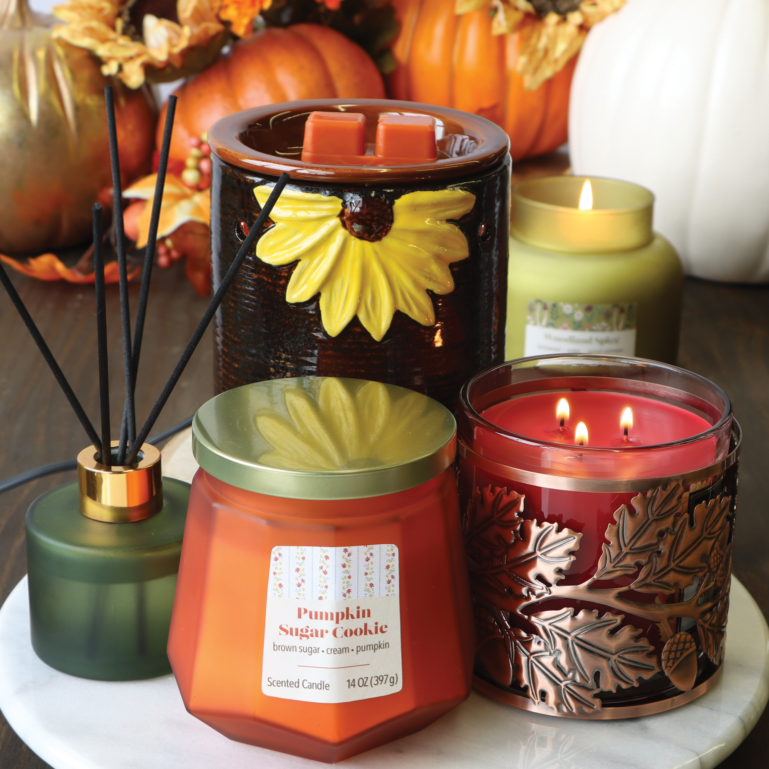 A variety of autumn-themed candles and diffusers are arranged on a white surface, featuring a Tuscany Candle® SEASONAL pumpkin sugar cookie candle, a red candle with leaf designs, and a Tuscany Candle® SEASONAL Sunflower Wax Melt Warmer that all emit delightful fragrances.