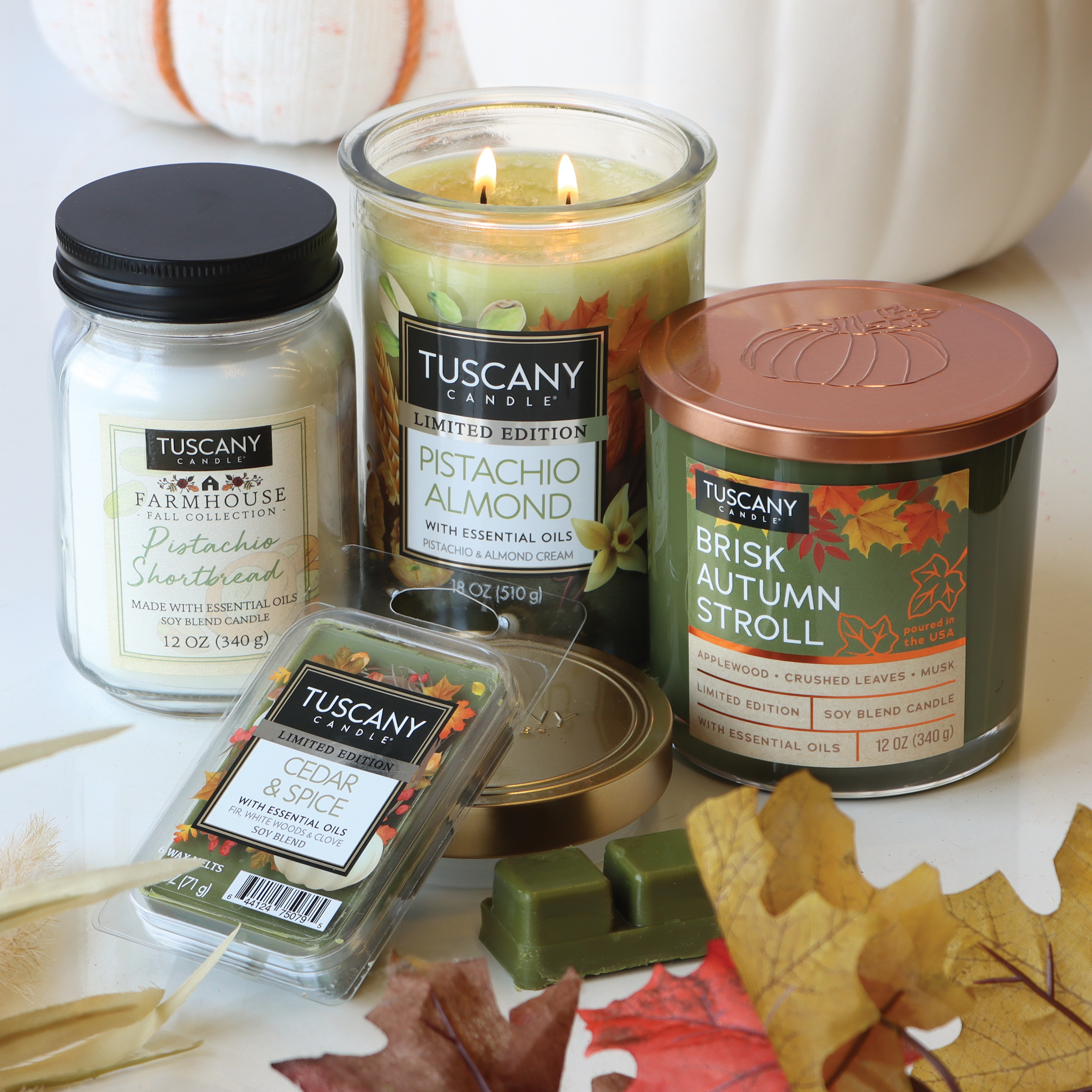 Four Tuscany Candle® SEASONAL candles in various scents, including a delectable Pistachio Almond Long-Lasting Scented Jar Candle (18 oz), Pistachio Shortbread, Brisk Autumn Stroll, and Cedar & Spice, arranged with autumn leaves in the background for the perfect Fall Collection Candle display.