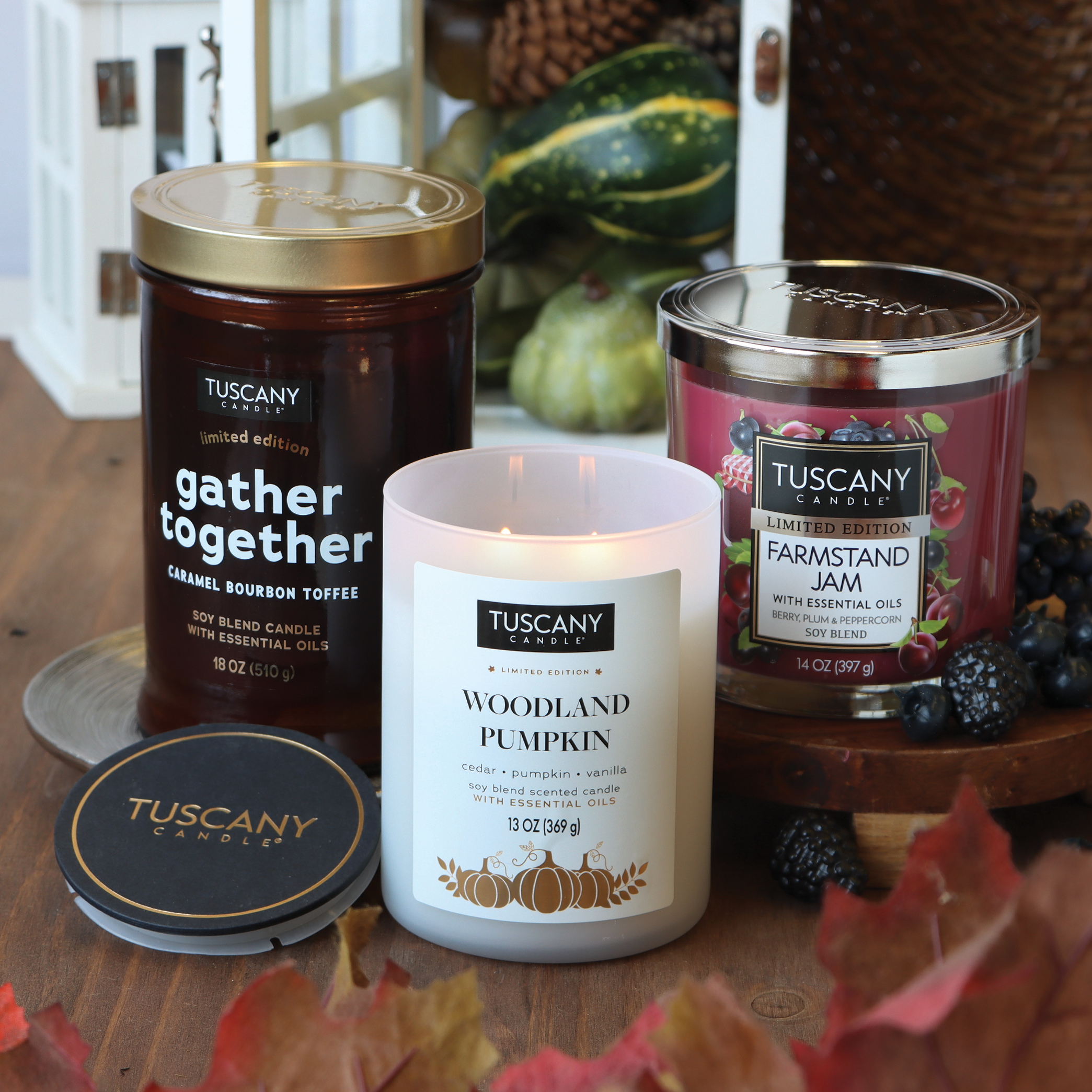 A wooden table displays three Tuscany candles from the Fall Collection: "Gather Together," "Woodland Pumpkin," and the fruity fragrance of the "Farmstand Jam Long-Lasting Scented Jar Candle (14 oz)." The decor includes a small artificial pumpkin, leaves, and berries.