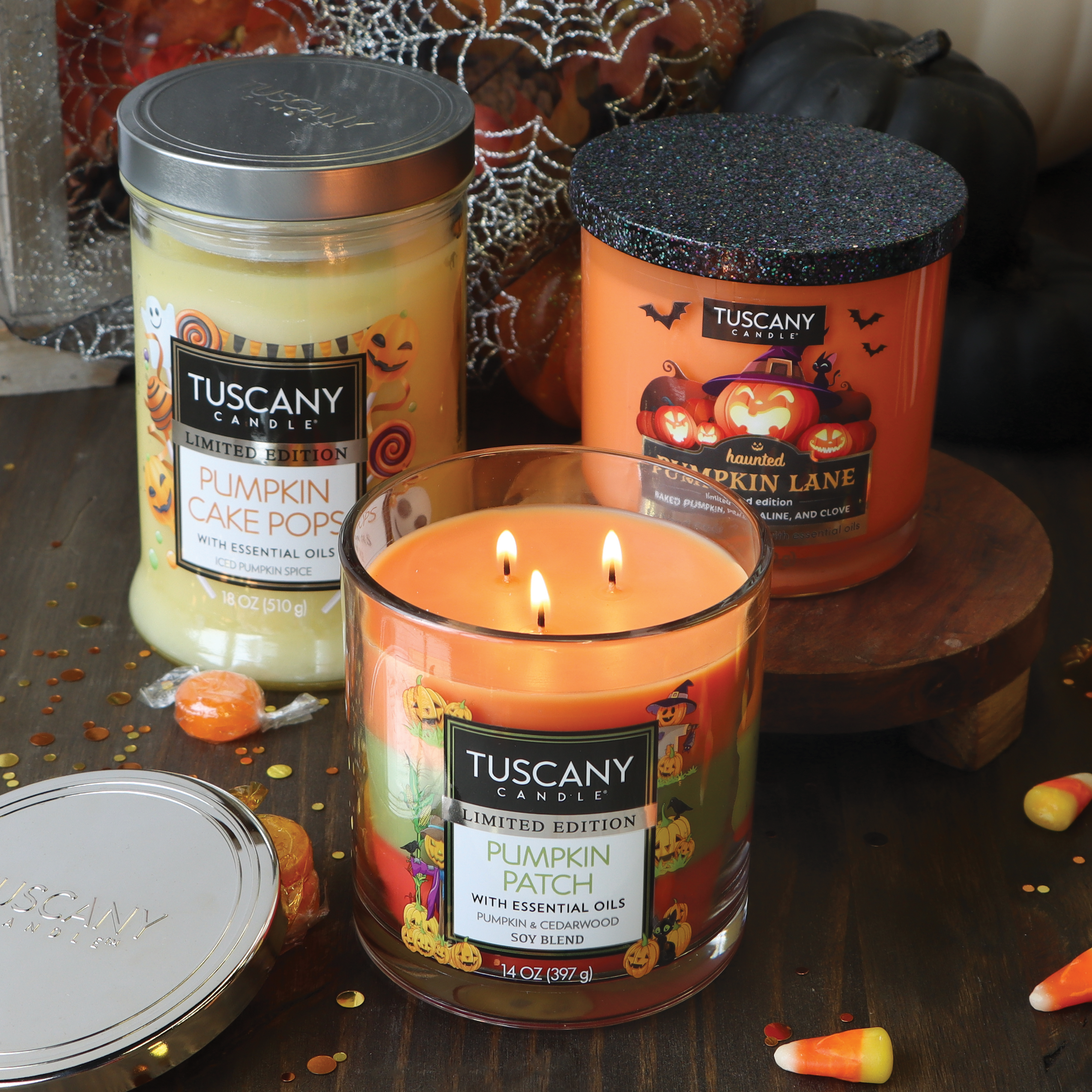 Displayed are three Tuscany 14 oz scented jar candles: "Pumpkin Cake Pops," "Haunted Pumpkin Lane," and "Pumpkin Patch Long-Lasting Scented Jar Candle." Additionally, candy corns are scattered around the candles, enhancing the cozy cedarwood aroma.