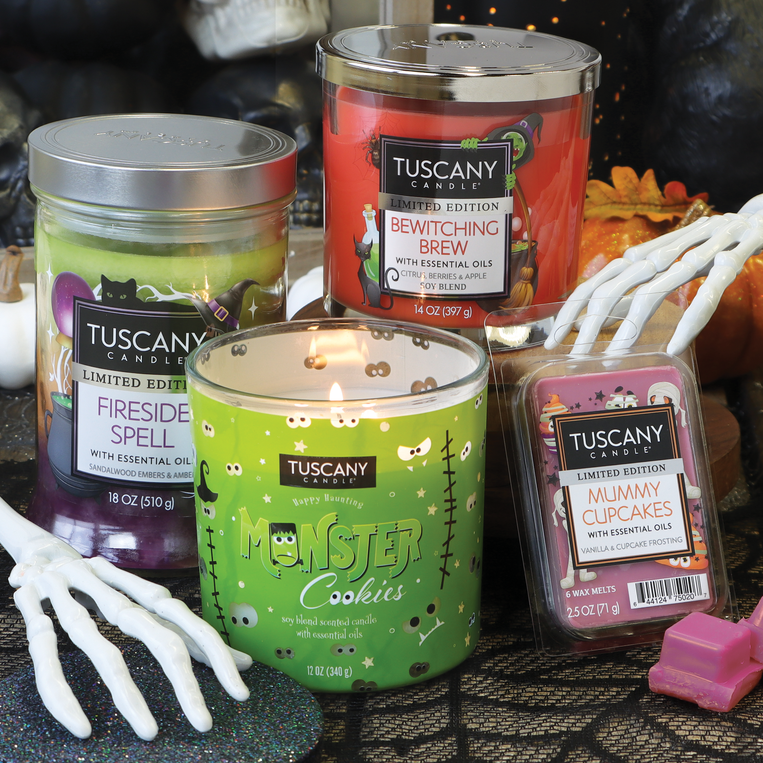 A display of Tuscany Candle® SEASONAL scented jar candles, including an "Fireside Spell" candle, a "Bewitching Brew" candle, a "Monster Cookies Long-Lasting Scented Jar Candle (12 oz)," and "Mummy Cupcakes" wax melts, set against a Halloween-themed backdrop as part of this limited-run collection.