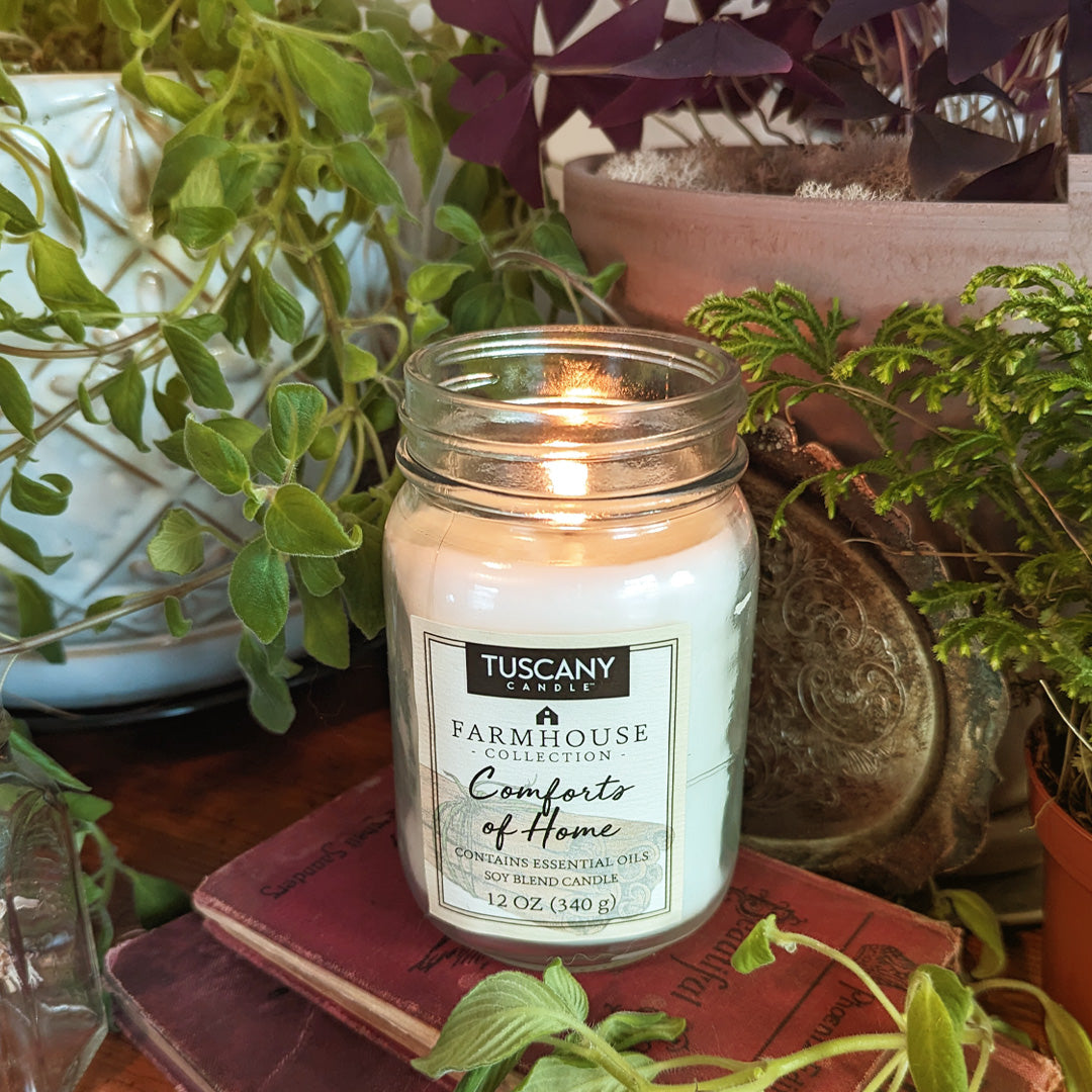 A jar of Comforts of Home Scented Jar Candle (12 oz) – Farmhouse Collection sits on a table next to books and plants, emitting a warm scent of cinnamon. The brand name is Tuscany Candle® EVD.
