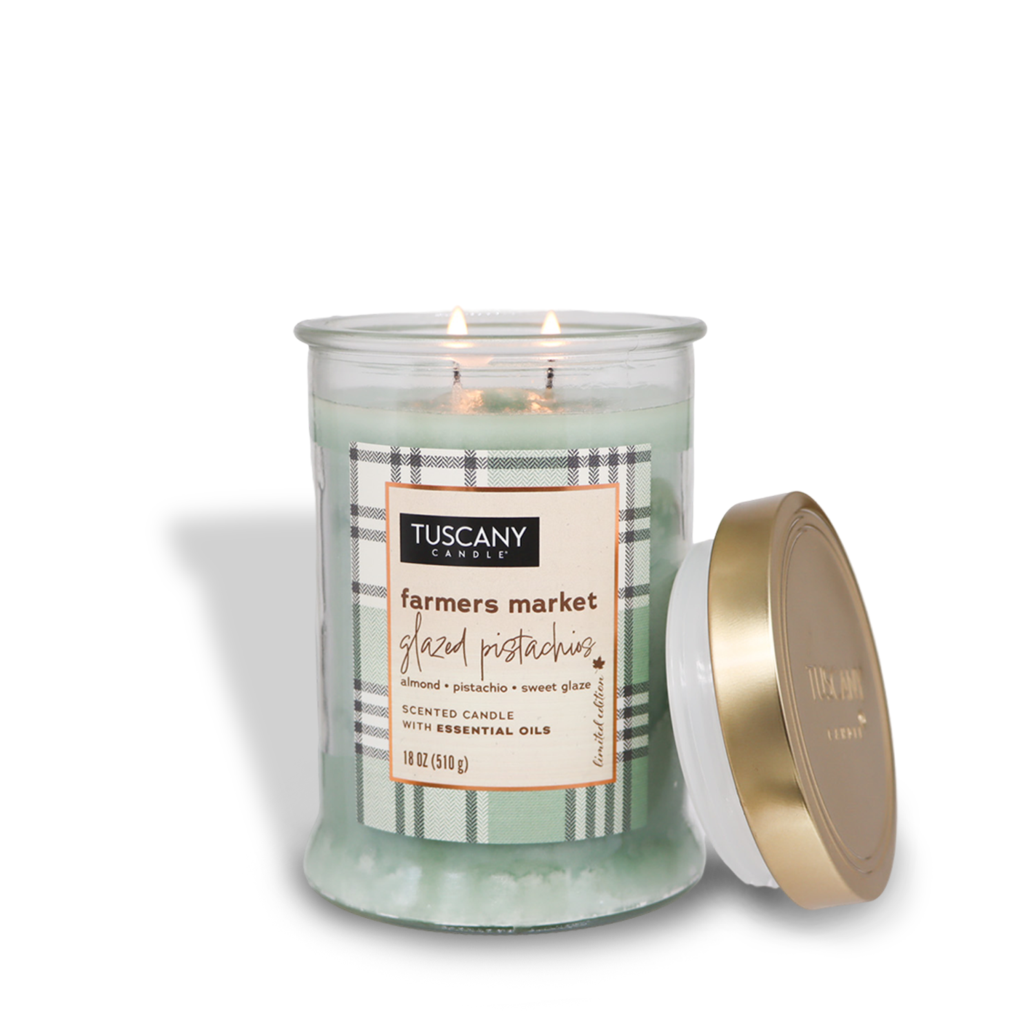 A lit Tuscany Candle® SEASONAL from the Autumn Flannels Collection in a glass jar labeled "Farmers Market Glazed Pistachios Long-Lasting Scented Jar Candle (18 oz)," featuring a gold lid placed beside it. The candle has light green wax inside.