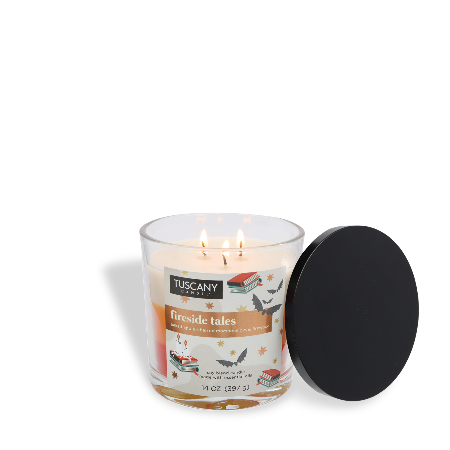 A lit 14 oz Tuscany Candle® SEASONAL glass jar candle, labeled "Fireside Tale: Charred Marshmallow & Baked Apple," with three wicks stands beside a black lid; the jar is decorated with book and bird illustrations.