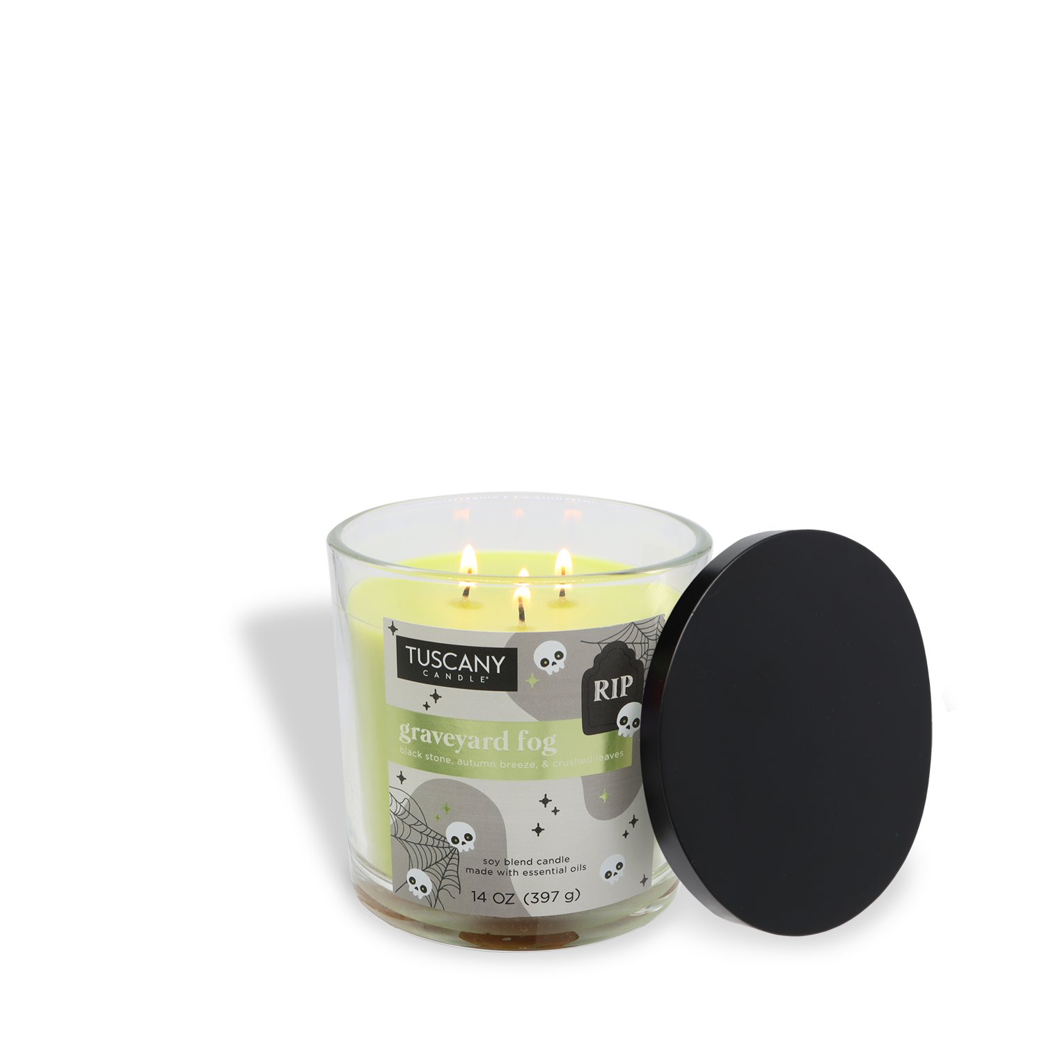 A 14 oz Tuscany Candle® SEASONAL “Graveyard Fog: Crushed Leaves & Autumn Breeze” Halloween candle, in a glass jar with three lit wicks, features skulls, spiderwebs, and festive graphics, plus a black lid beside the jar.