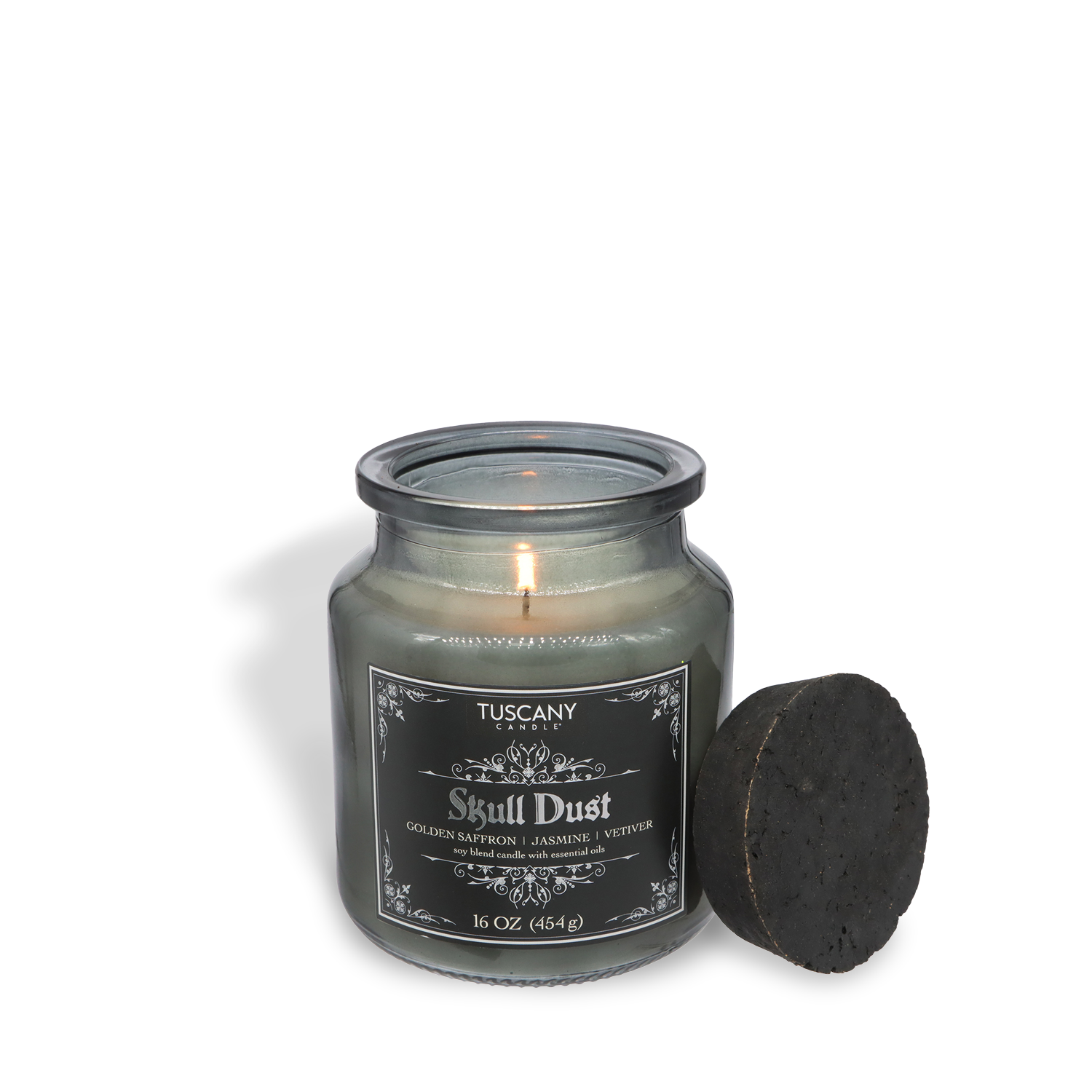 A lit Tuscany Candle® SEASONAL Skull Dust: Fireside Vanilla & Smooth Vetiver candle (16 oz) in an apothecary jar, black lid beside it, sets a mysterious Halloween mood with its rich seasonal fragrance.