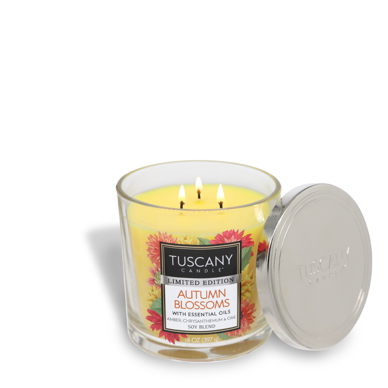 A lit yellow Tuscany Candle® SEASONAL labeled "Autumn Blossoms Long-Lasting Scented Jar Candle (14 oz)" with three wicks, featuring fragrance notes of amber, chestnut, and oak from its premium satin wax, next to a silver lid.