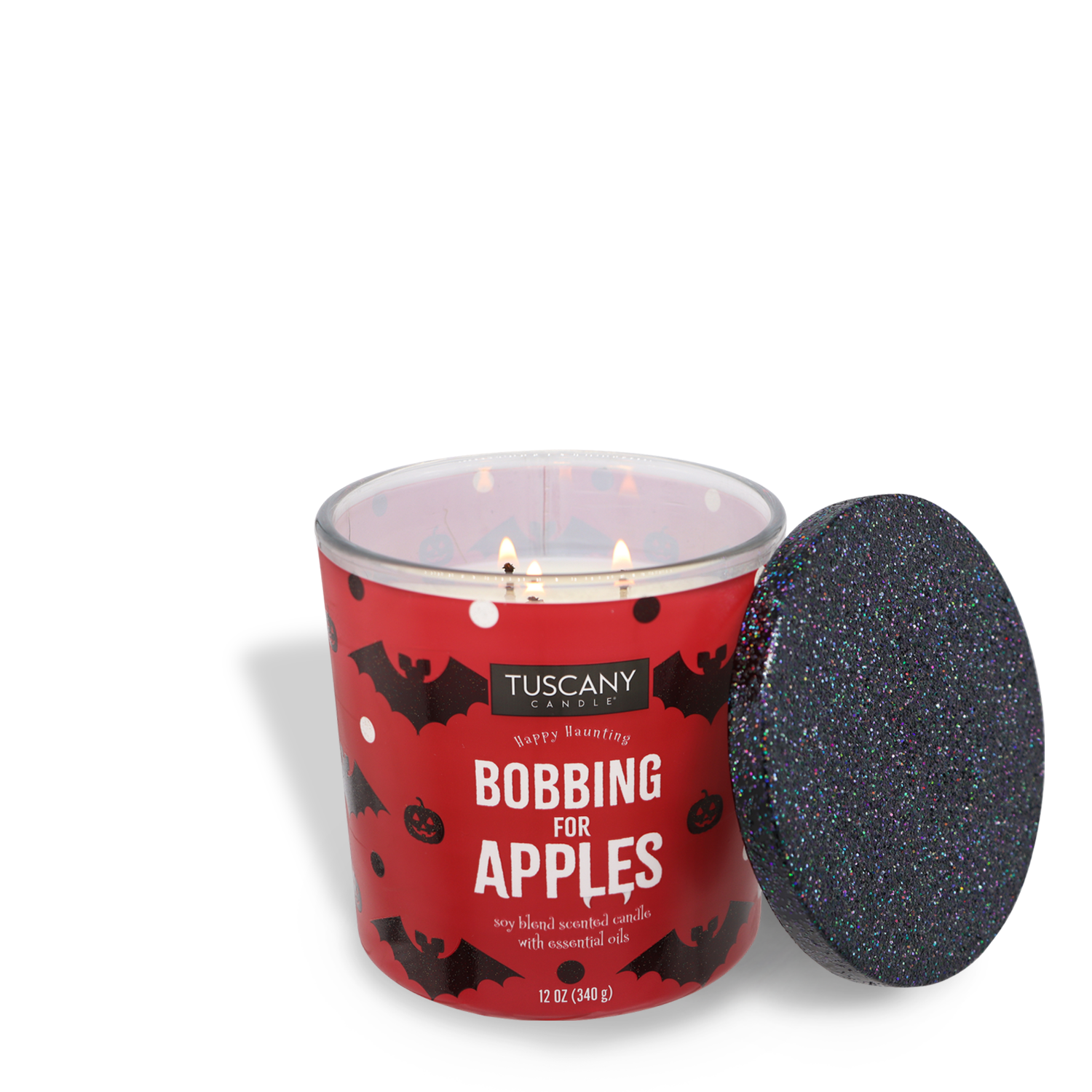 A lit scented jar candle with the label "Tuscany Candle® SEASONAL, Bobbing For Apples Long-Lasting Scented Jar Candle (12 oz)" sits in a red container decorated with bat and apple designs. Made of premium satin wax, its fragrance notes create a cozy atmosphere. The lid is removed and placed to the side.