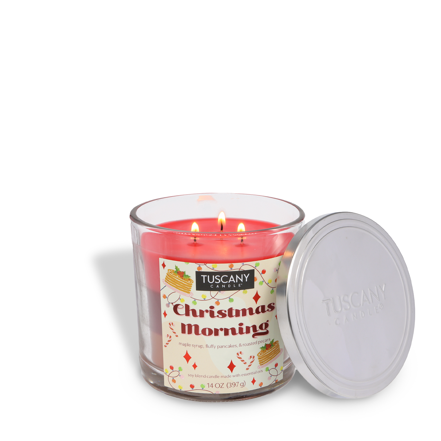 A lit Tuscany Candle® SEASONAL Christmas Morning Scented Holiday Candle (14 oz) in a glass jar with a silver lid beside it. The label features notes of maple syrup, fluffy pancakes, and cinnamon.