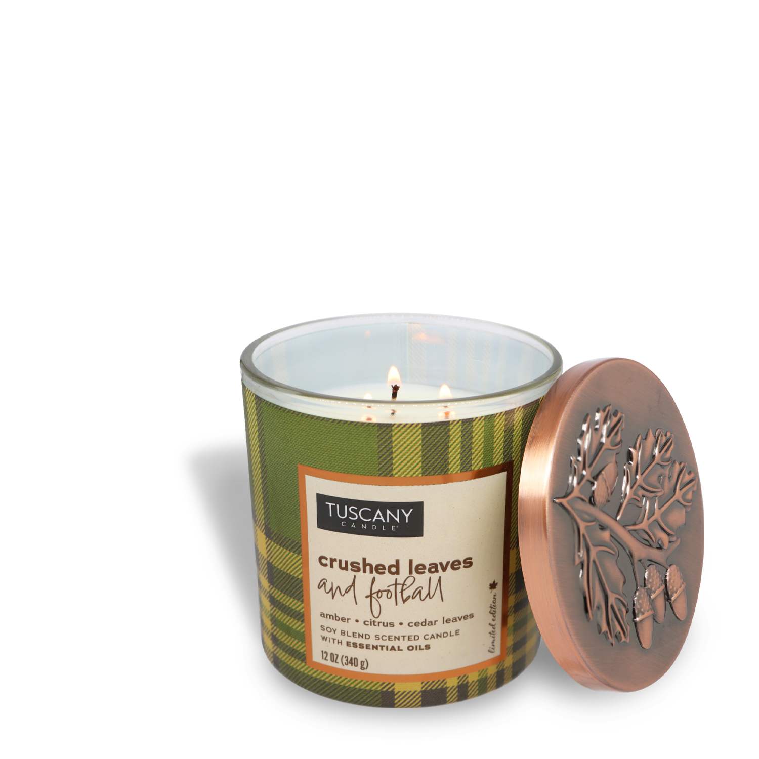 A Tuscany Candle® SEASONAL "Crushed Leaves and Football Long-Lasting Scented Jar Candle (12 oz) — Autumn Flannels Collection," housed in a green plaid-patterned jar with an embossed tree design on the lid, sits with the lid off to the side. The three lit wicks of the candle fill the room with its warming fragrance.