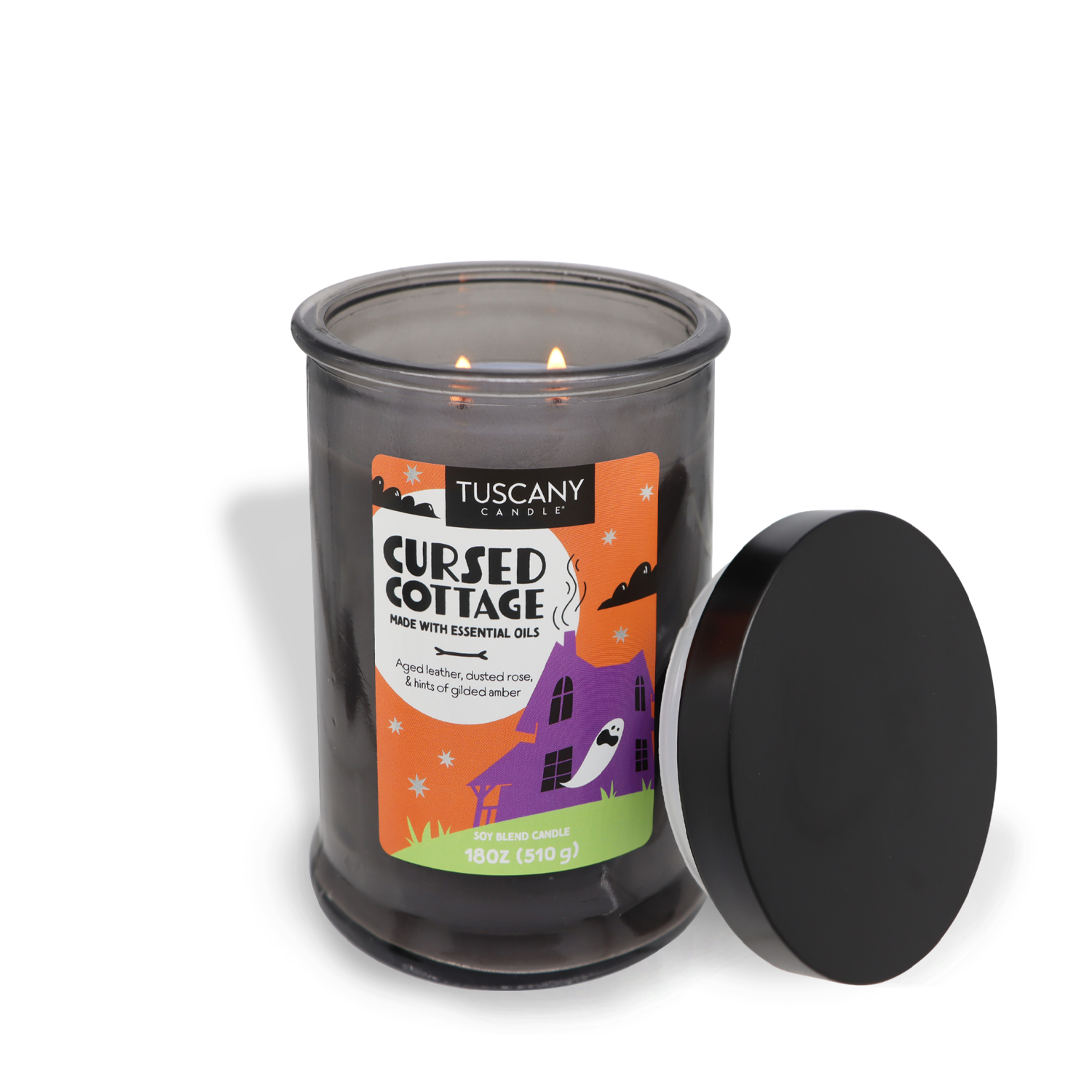 A lit "Cursed Cottage Long-Lasting Scented Jar Candle (18 oz)" from Tuscany Candle® SEASONAL rests in a glass jar with an illustrated label of a haunted house. The candle lid is placed on its side next to the jar, releasing the enchanting aroma of essential oils into the room.