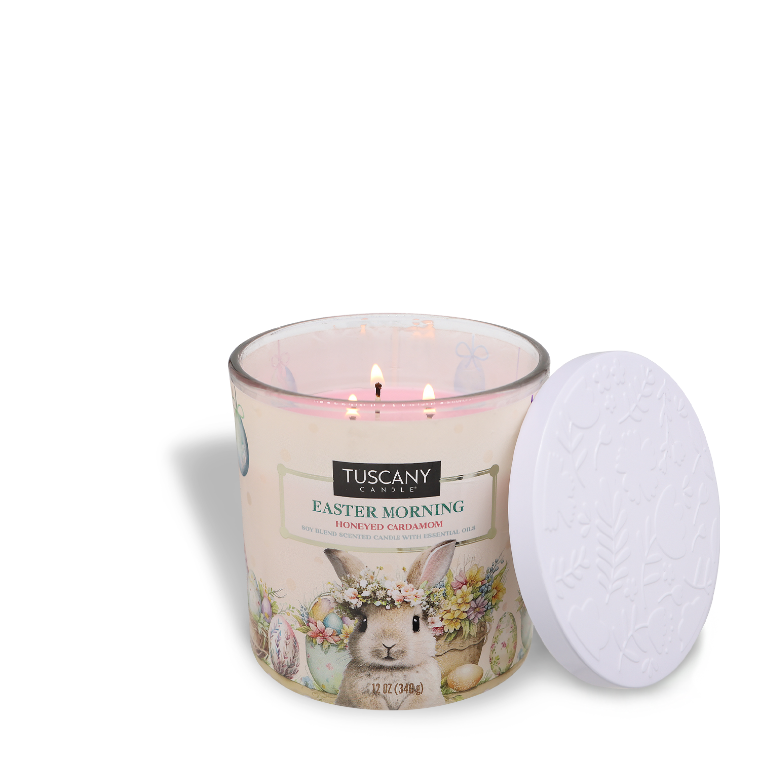 A lit three-wick candle, "Easter Morning: Cardamom & Honey," by Tuscany Candle® SEASONAL, with a floral and bunny design and a white embossed lid beside it.