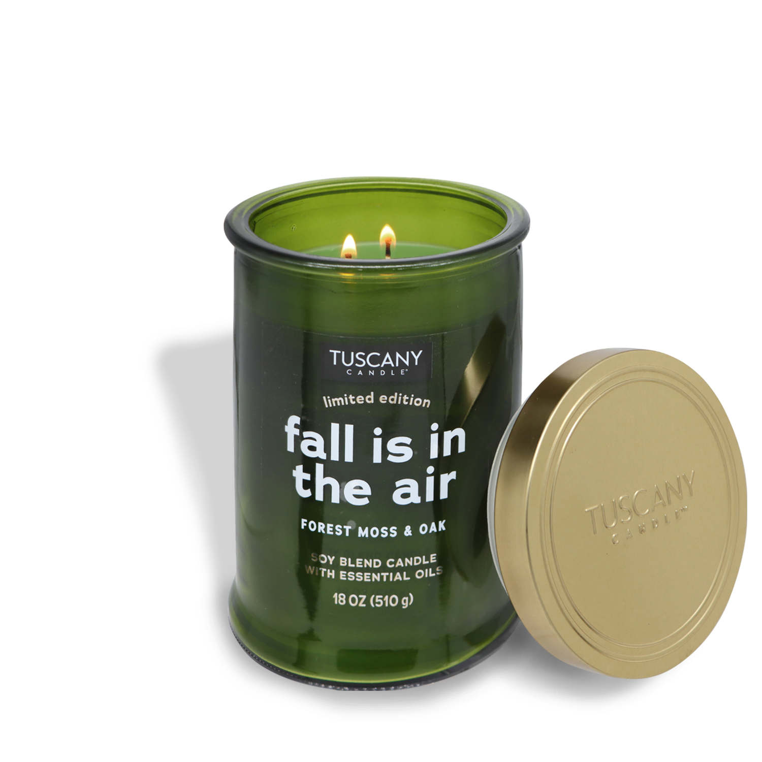 An 18 oz (510 g) "Fall is in the Air" long-lasting scented jar candle from the Tuscany Candle® SEASONAL Gather Together Collection, with a forest moss & oak autumn fragrance, sits lit and emitting a green glow next to its detached gold lid. This beautifully crafted soy blend fall candle encapsulates the essence of autumn.