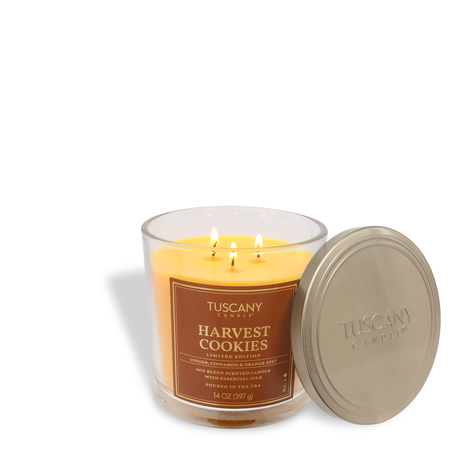 A 14 oz "Harvest Cookies: Brown Sugar & Vanilla" candle by Tuscany Candle® SEASONAL with three lit wicks sits beside its metallic embossed lid.