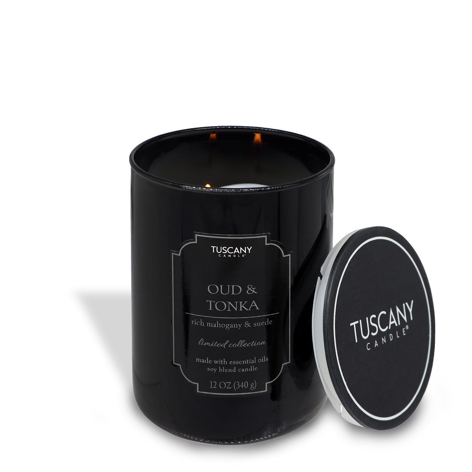 A lit Oud & Tonka (12 oz) candle in a black glass container, labeled "Mahogany Suede" with a metal lid next to it, on a white background.
