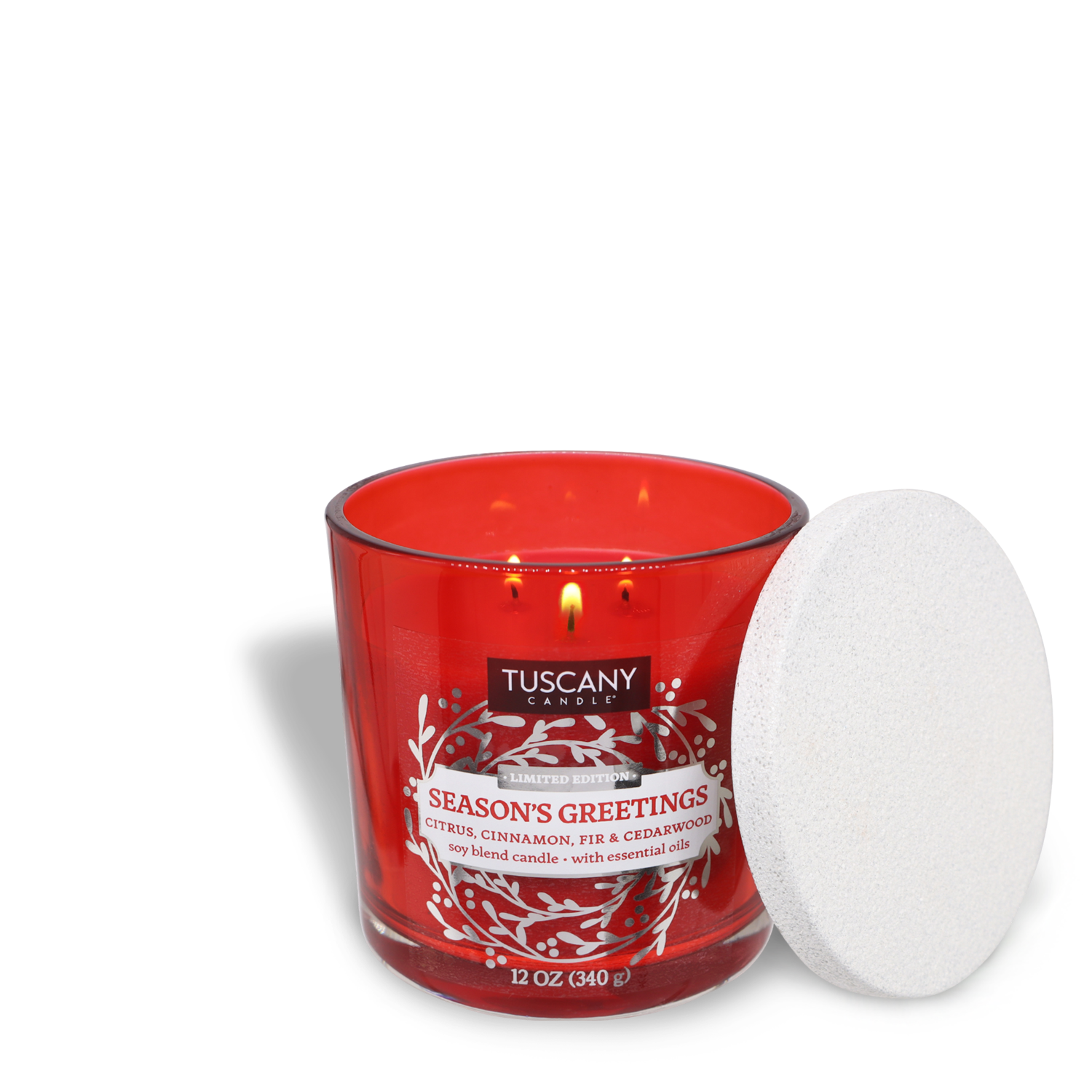 A 12 oz red Tuscany Candle® SEASONAL jar candle labeled "Season's Greetings," featuring a citrus, cinnamon, fir, and cedarwood scent. The candle has three lit wicks and a white lid placed beside it.