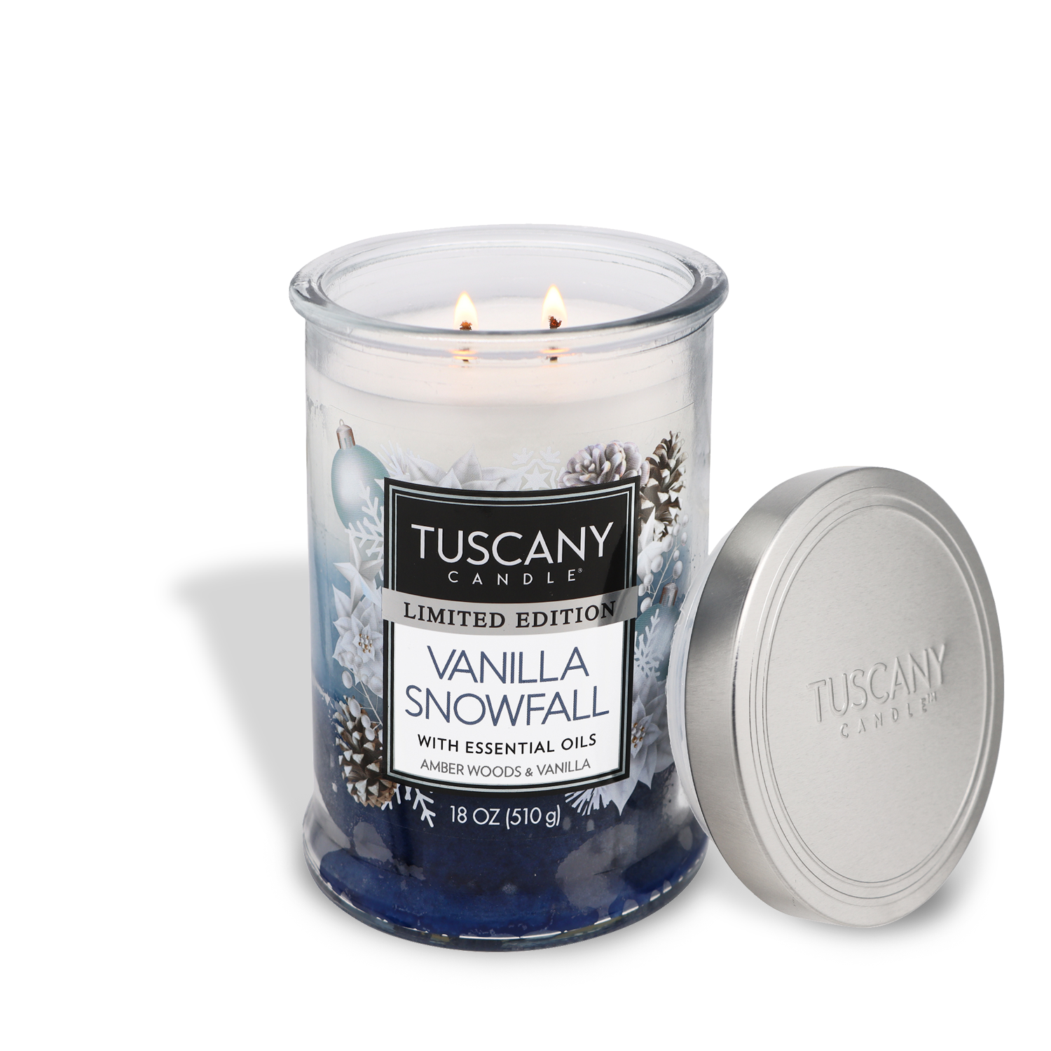 A lit Tuscany Candle® SEASONAL Vanilla Snowfall Long-Lasting Scented Jar Candle, made with essential oils in warm amber woods and sweetened vanilla, presented in a glass jar with a lid beside it. The candle is an 18 oz limited edition.