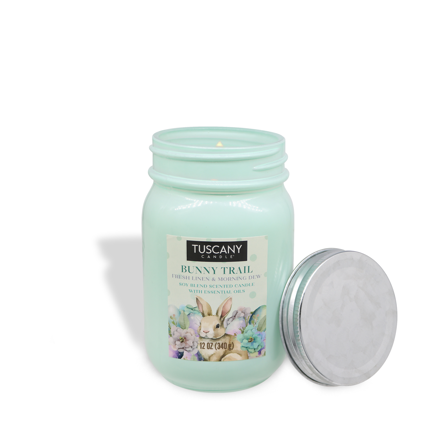 A light green "Bunny Trail" candle jar by Tuscany Candle® SEASONAL, featuring rabbit and flower designs, has its lid placed beside it. It is a 12 oz linen-scented Easter candle.