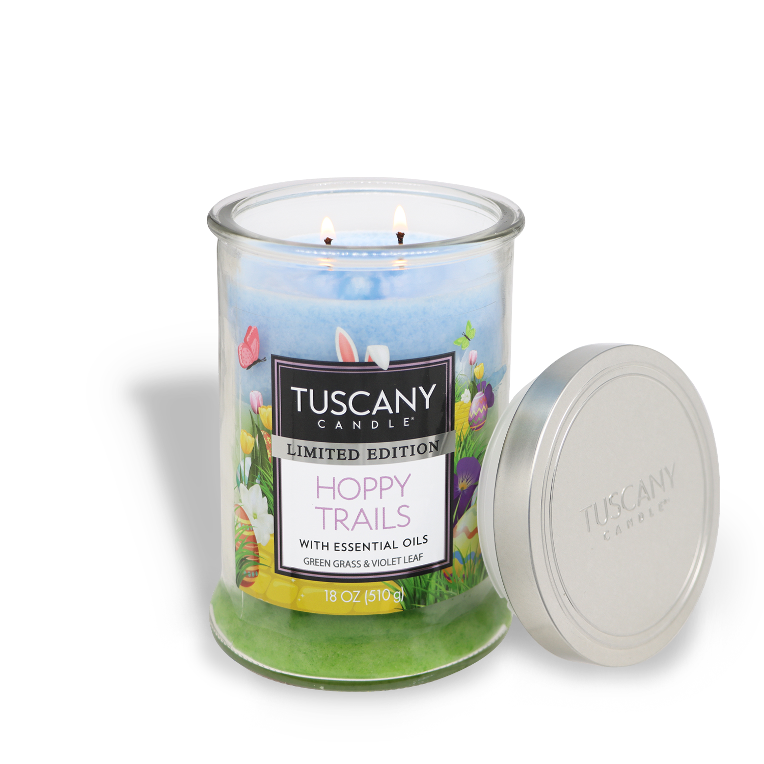 An open clear glass jar candle with three burning wicks labeled "Tuscany Candle® SEASONAL, Hoppy Trails: Grass & Violet Scented Easter Candle (18 oz)" features a spring-themed design.