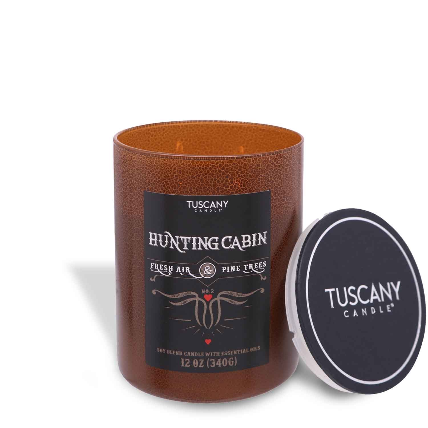 A Tuscany Candle® SEASONAL Hunting Cabin Scented Jar Candle (12 oz) – Rugged Retreat Collection with a lid next to it, emitting the fresh aroma of juniper berry and pine woods.