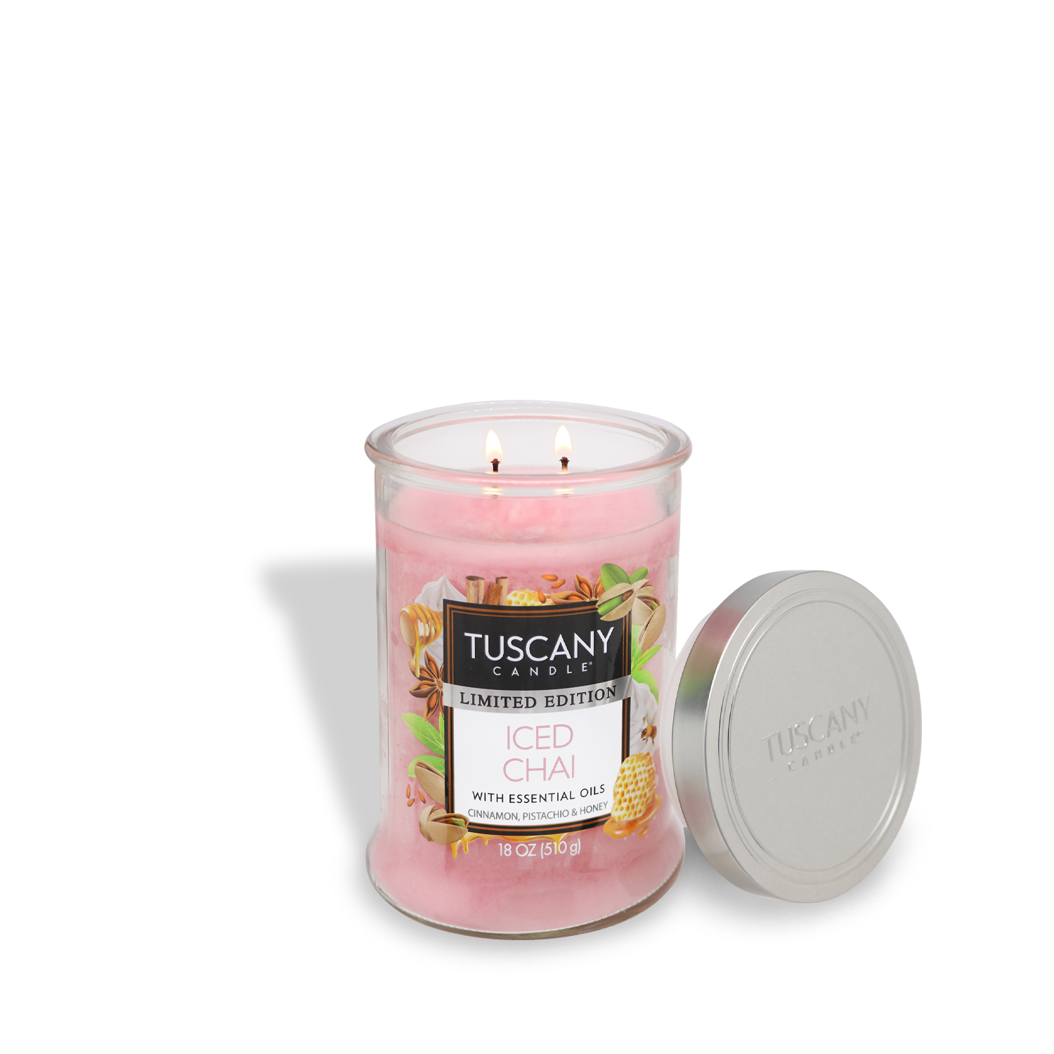 A pink wax glass jar candle labeled "Tuscany Candle® SEASONAL Iced Chai," featuring an 18 oz cardamom and honey scent with two wicks, is displayed with its metal lid beside it.