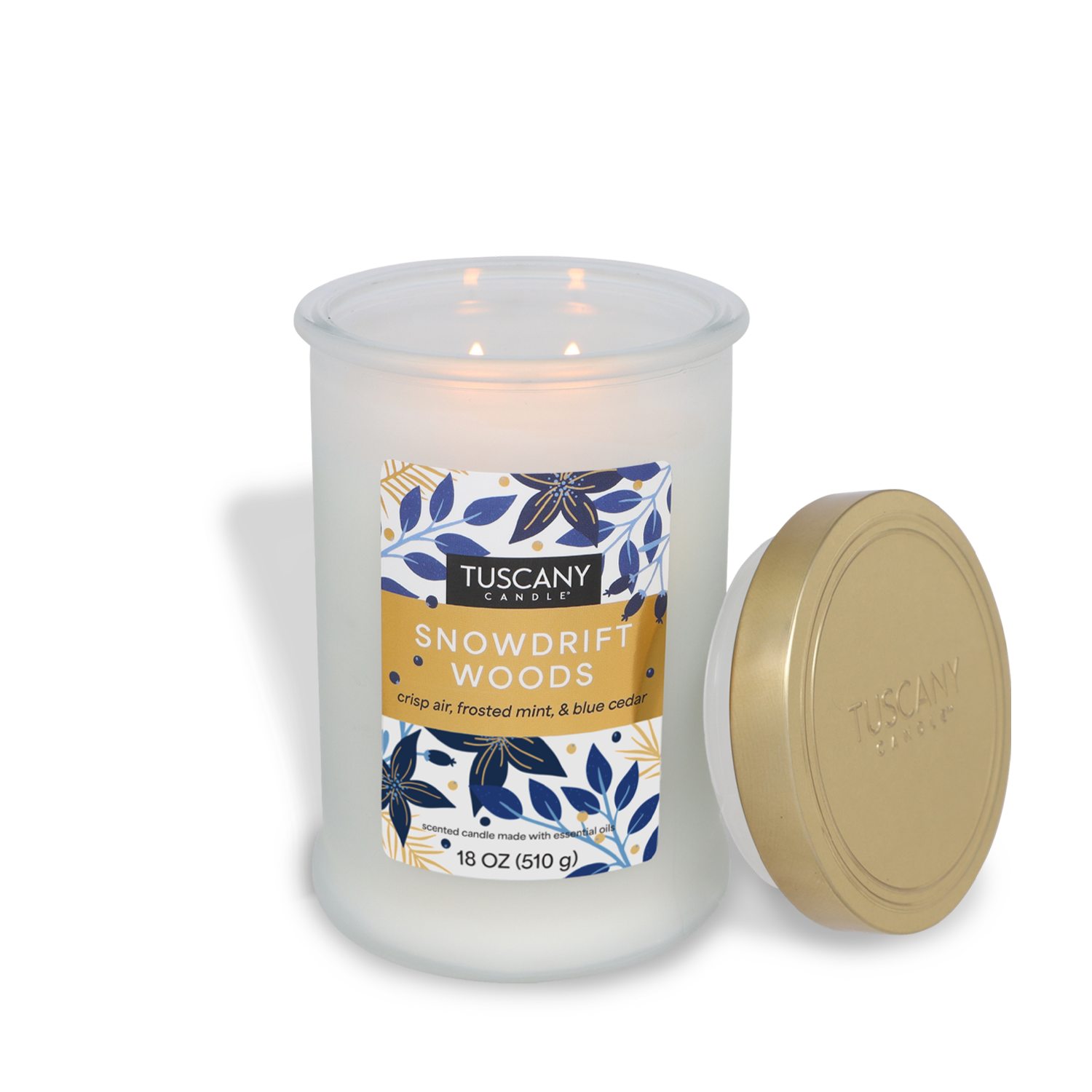 A lit Tuscany Candle® SEASONAL "Snowdrift Woods" scented jar candle (18 oz) emitting fragrances of crisp air, frosted mint, and blue cedar, with its gold lid resting beside it.