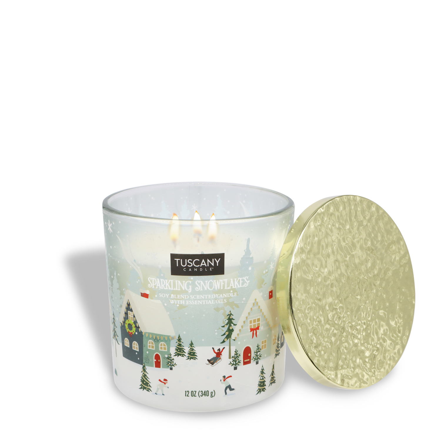 A lit Sparkling Snowflakes Scented Jar Candle (12 oz) by Tuscany Candle® SEASONAL, featuring a festive winter village design on the glass jar with a detached gold lid beside it.