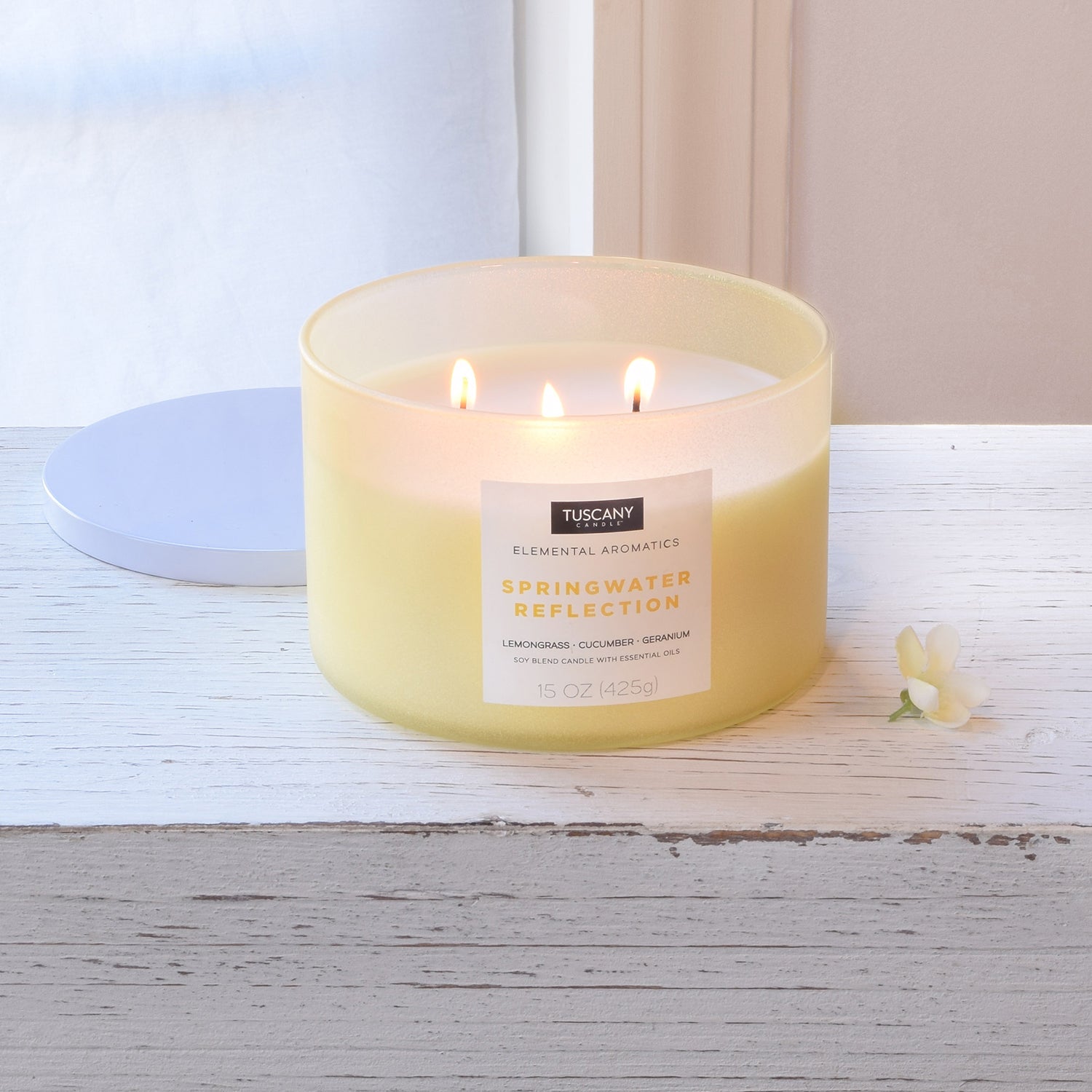 A Tuscany Candle Springwater Reflection Scented Jar Candle (15 oz) – Elemental Aromatics Collection sits on a table next to a window.