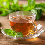 Load image into Gallery viewer, A photo of a glass teacup surrounded by mint leaves. The image represents the two main olfactory notes in the &quot;tea Leaves&quot; scented candle from Tuscany Candle.

