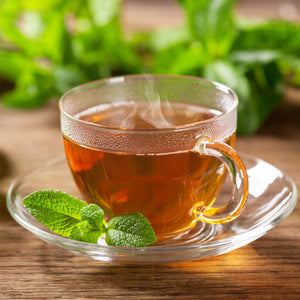 A photo of a glass teacup surrounded by mint leaves. The image represents the two main olfactory notes in the "tea Leaves" scented candle from Tuscany Candle.