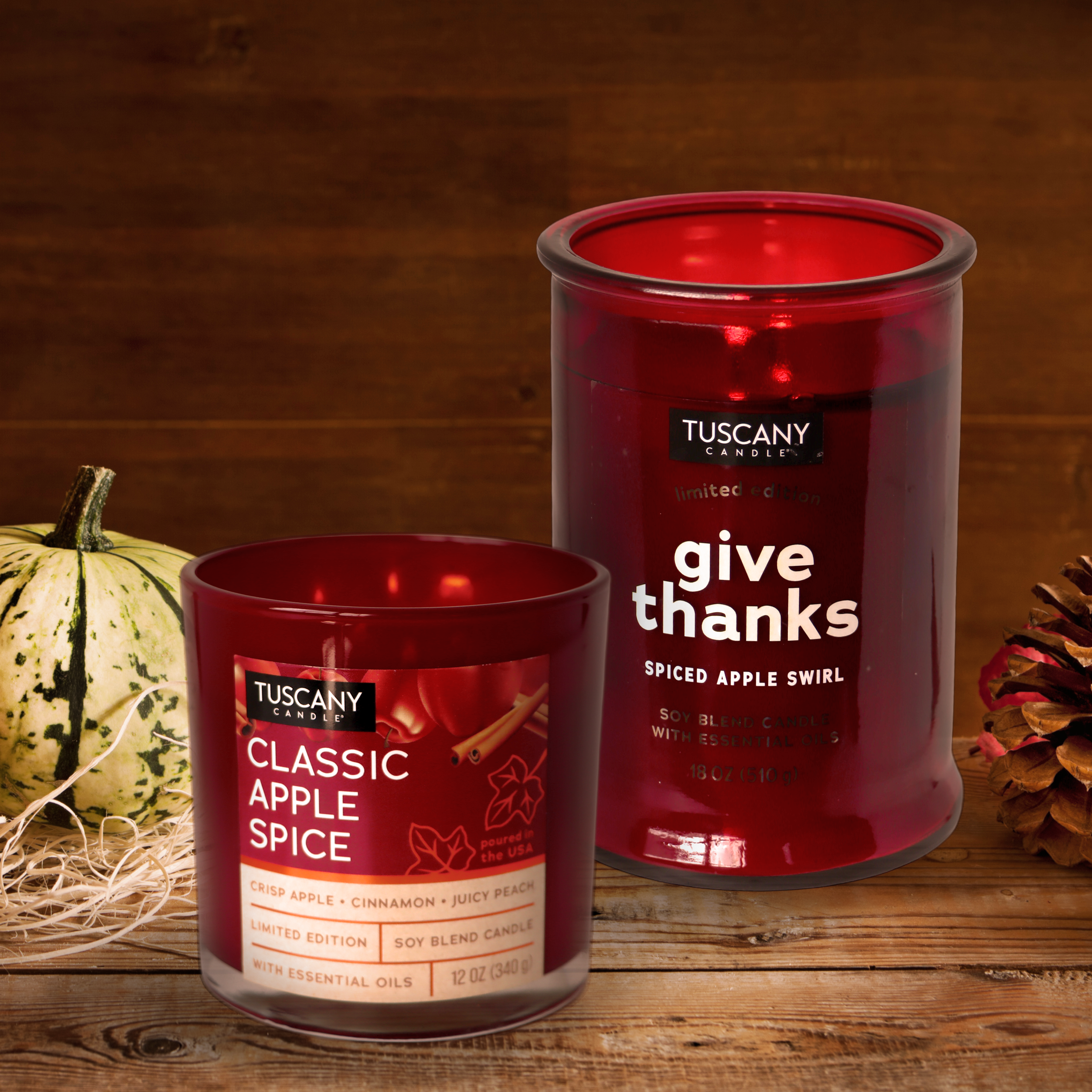 Two Tuscany Candle® SEASONAL candles are displayed. The front candle, from the Copper Harvest Collection, is labeled "Classic Apple Spice Long-Lasting Scented Jar Candle (12 oz)," and the back candle is labeled "Give Thanks." A small pumpkin and an autumn leaf decoration are nearby, enhancing the autumn fragrance.