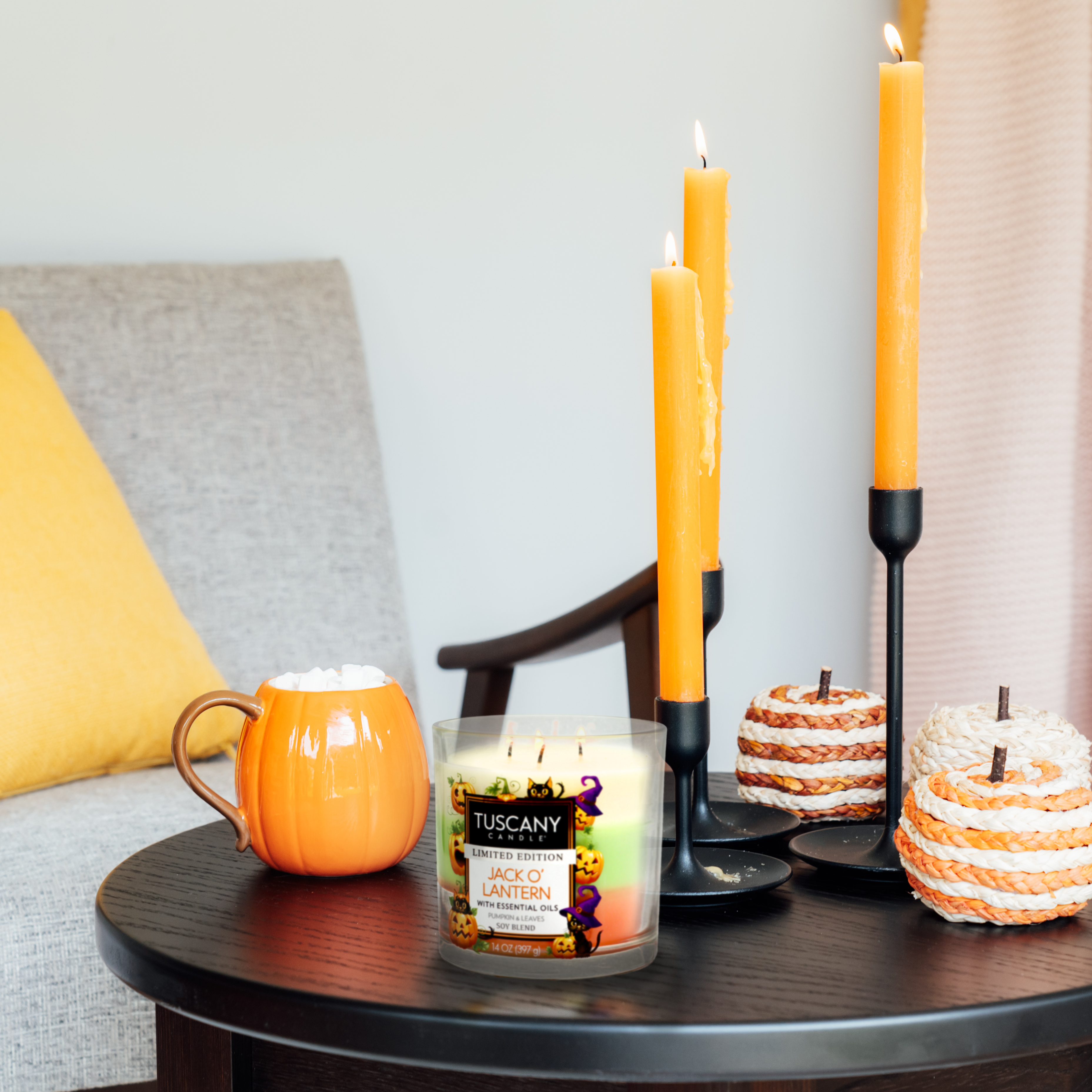 A small table adorned with lit orange candles, a Jack O' Lantern Long-Lasting Scented Jar Candle (14 oz) by Tuscany Candle® SEASONAL, a pumpkin-shaped mug filled with a drink, and two knitted pumpkins creates a Halloween vibe. An armchair with a yellow pillow sits in the background, perfect for enjoying scented jar candles.