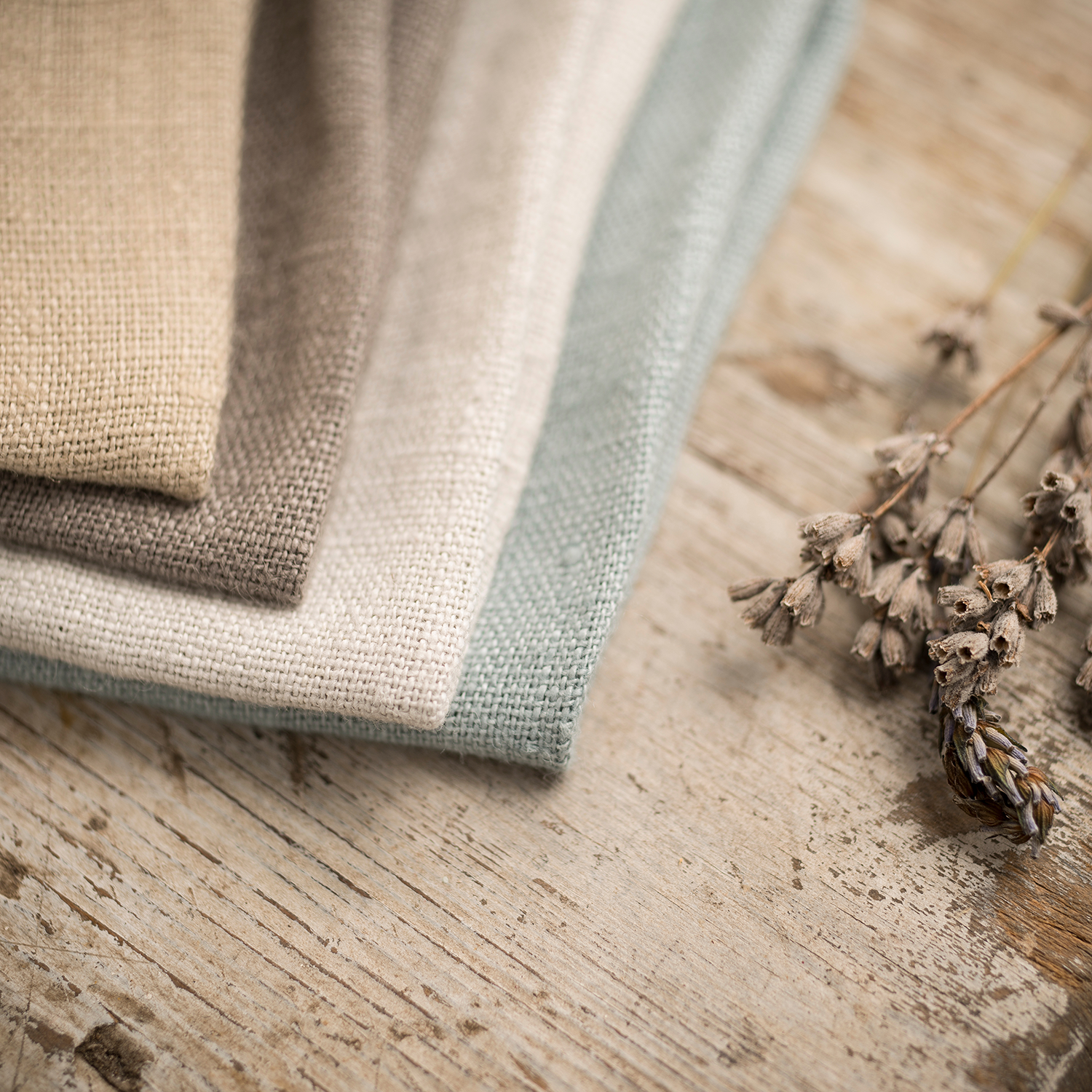 Three folded Belgium Linen (12 oz) napkins from the Colorsplash collection by Tuscany Candle® EVD beside dried flowers on a rustic wooden surface.