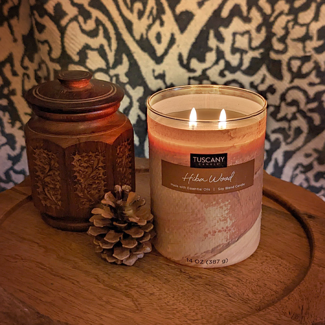 A Tuscany Candle Hiba Wood Scented Jar Candle (14 oz) – Home Décor Collection sits on a wooden table next to a pine cone.
