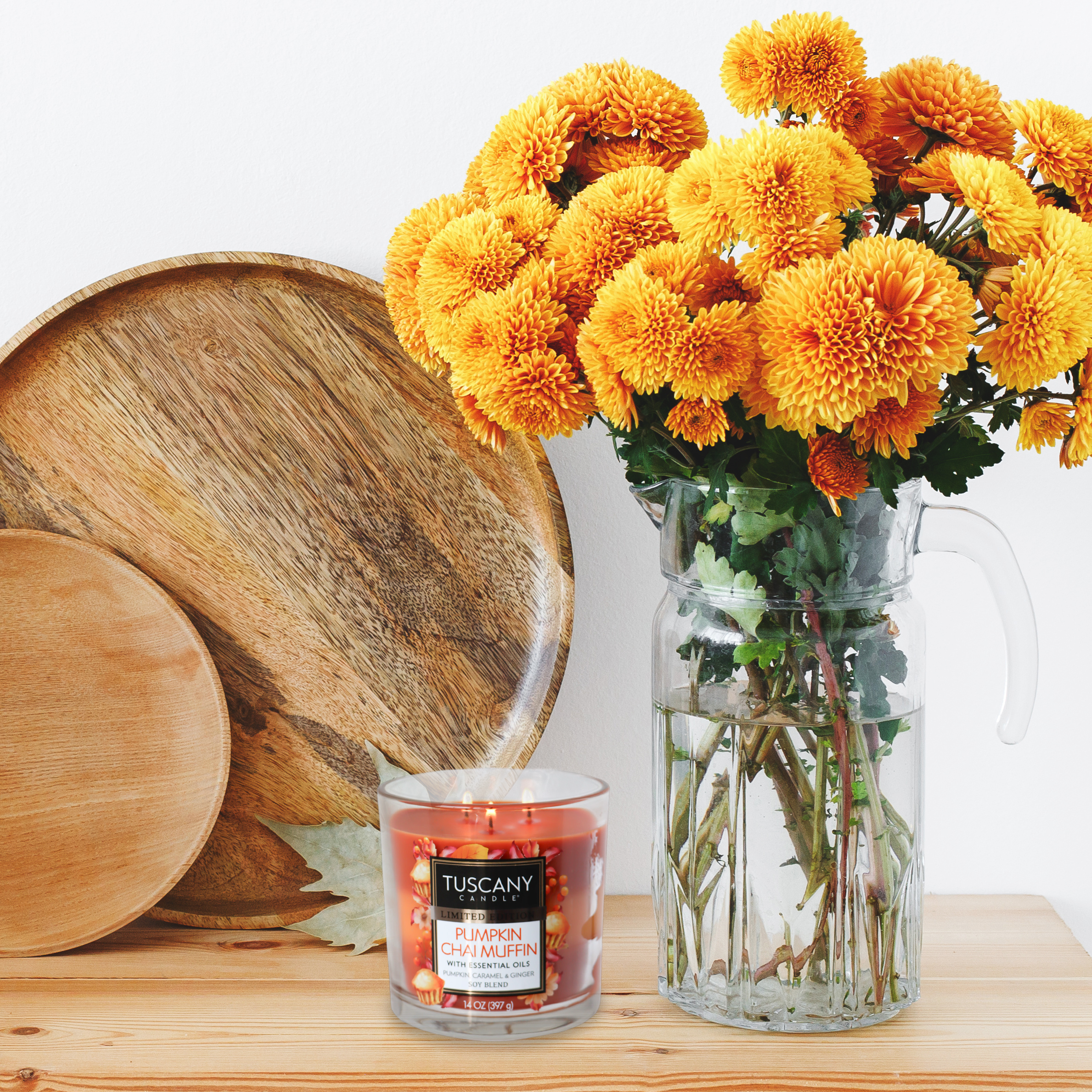 A lit Pumpkin Chai Muffin Long-Lasting Scented Jar Candle (14 oz) by Tuscany Candle® SEASONAL and a glass pitcher filled with bright orange chrysanthemums sit on a table beside wooden plates, making it a perfect addition to any fall collection.