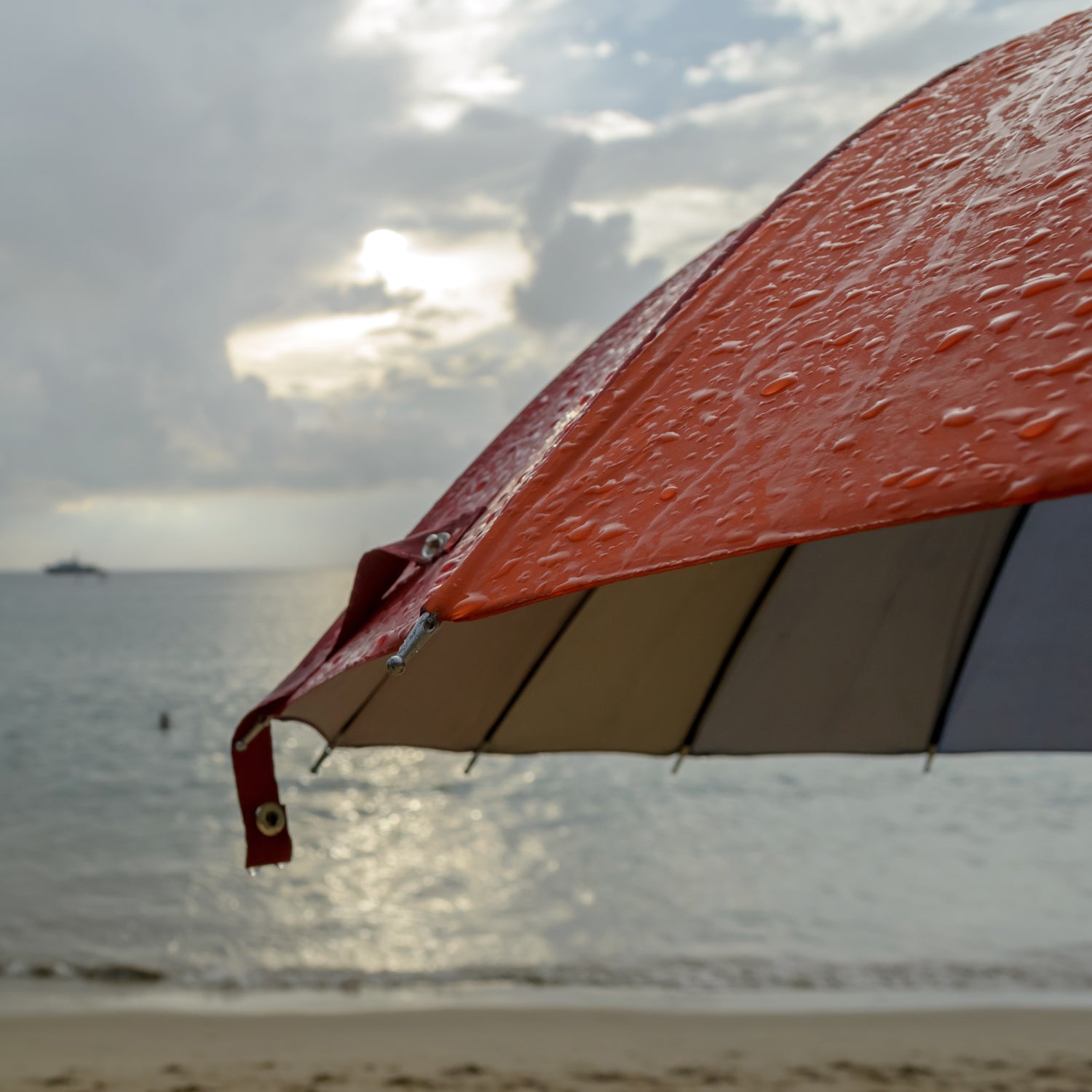 A red and orange Tuscany Candle® EVD umbrella on a beach, providing shade and protection from the sun while trying to catch the soothing scent of Sea & Sand Long-Lasting Scented Jar Candles (18 oz) wafting through the breeze.